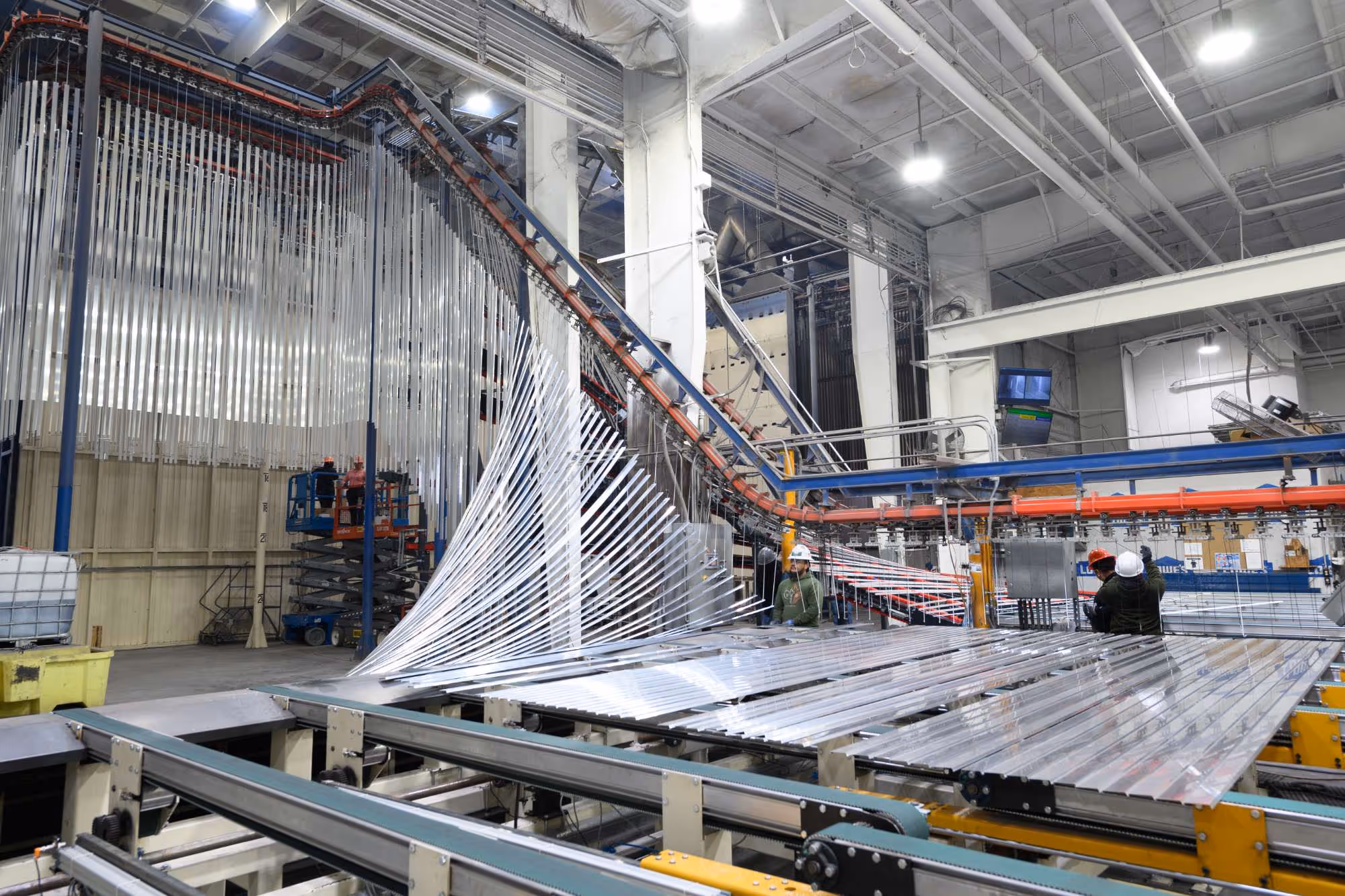 Workers wearing hard hats operating machinery in a large factory with overhead rails and suspended metal strips.