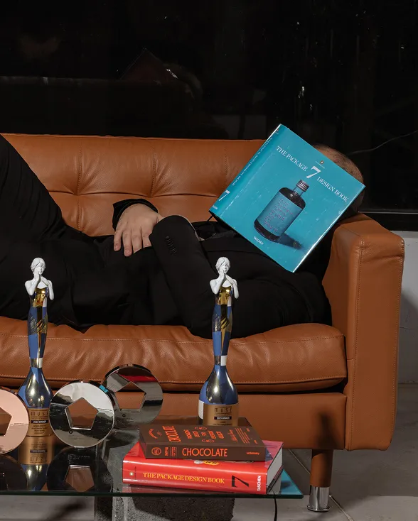 A man dressed in black lies on a brown sofa with a turquoise-covered book covering his face, next to several trophies and books on a table in front.
