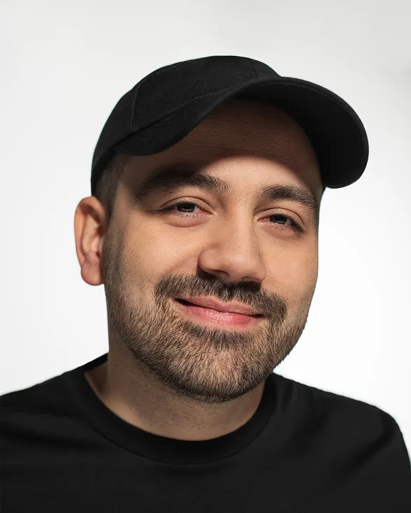 A man with a beard and a black hat smiling against a light background.