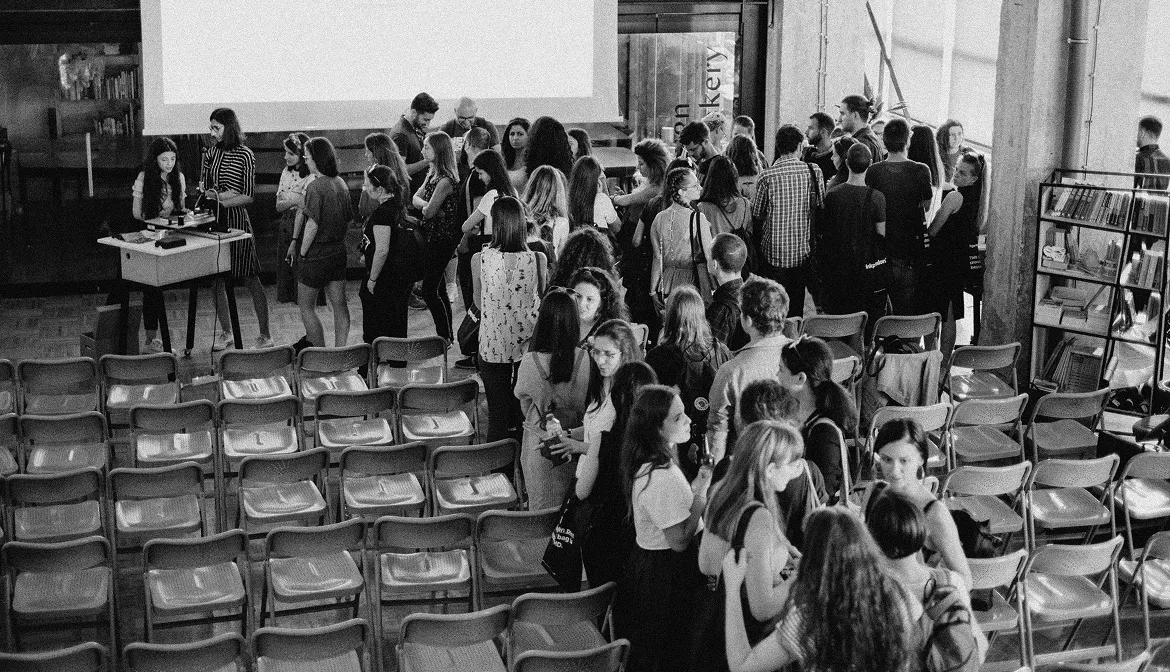 A group of people in conversation stand between empty rows of folding chairs in a hall.