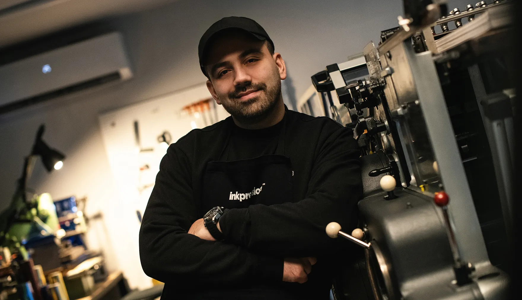 A man in a black hat and apron leaning on a machine in a workshop.