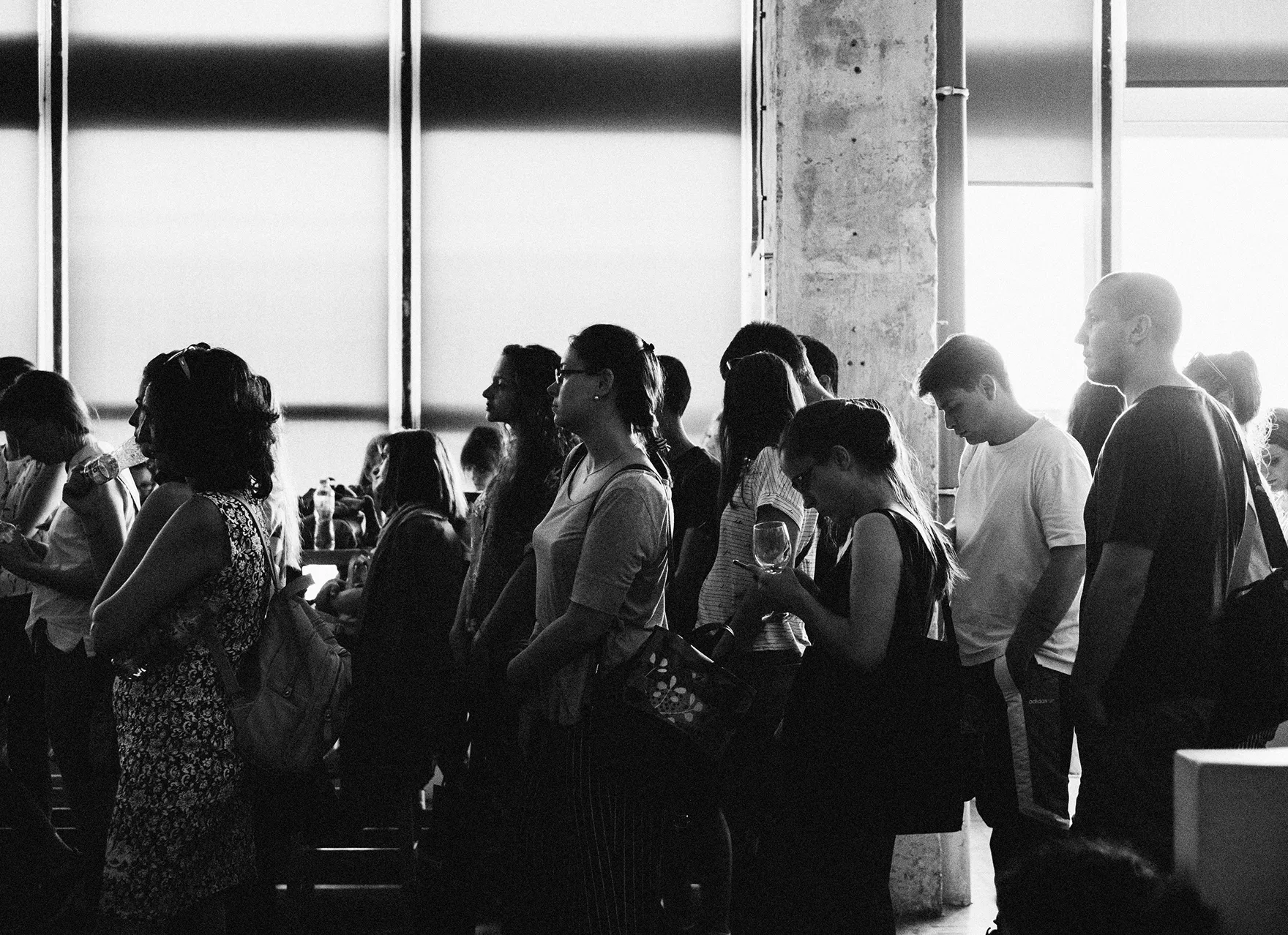A group of people standing in line inside a building with bright sunlight through large windows.