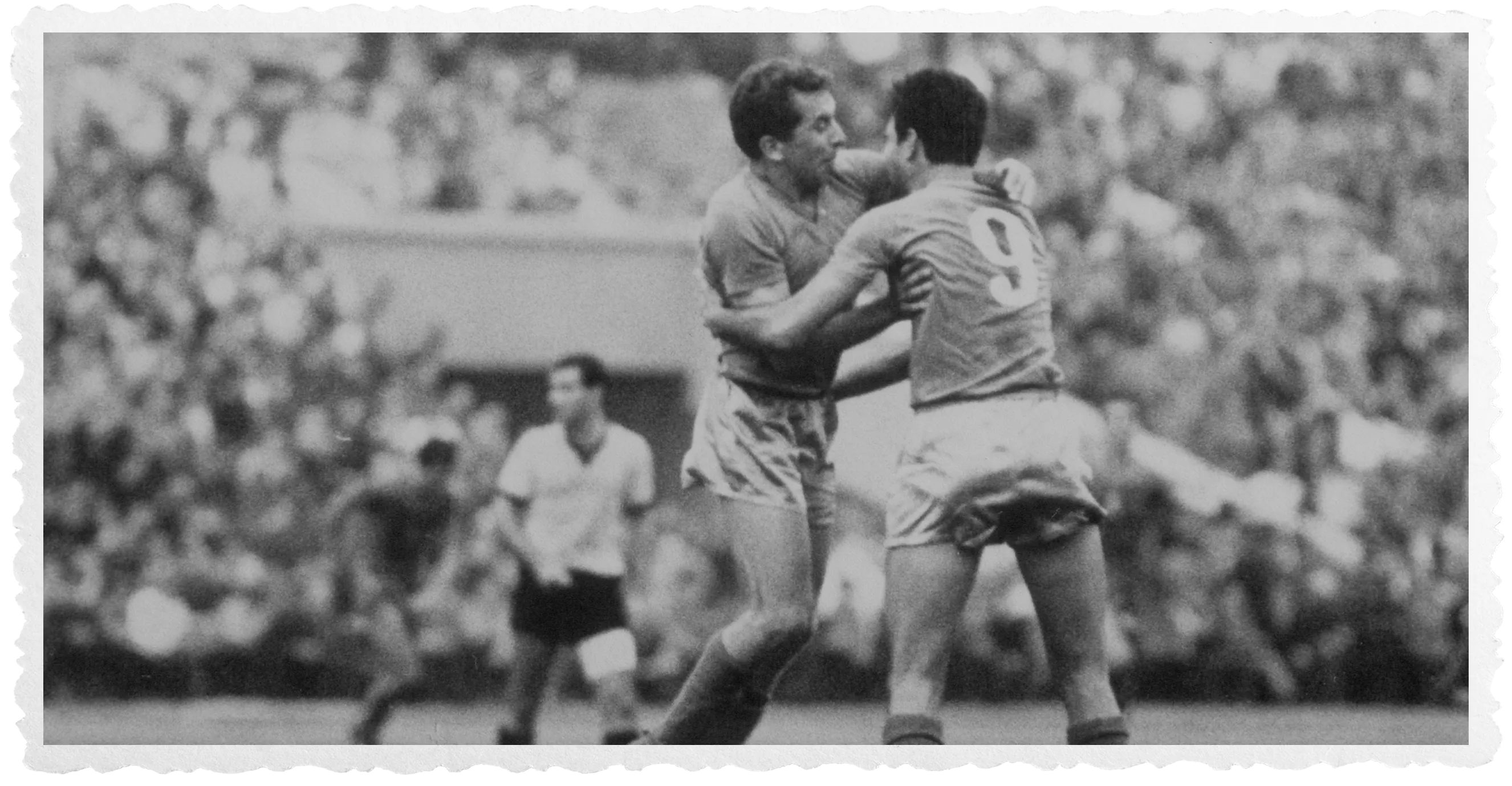 A black and white photo of two football players embracing during a match in a stadium full of spectators, possibly celebrating a goal.