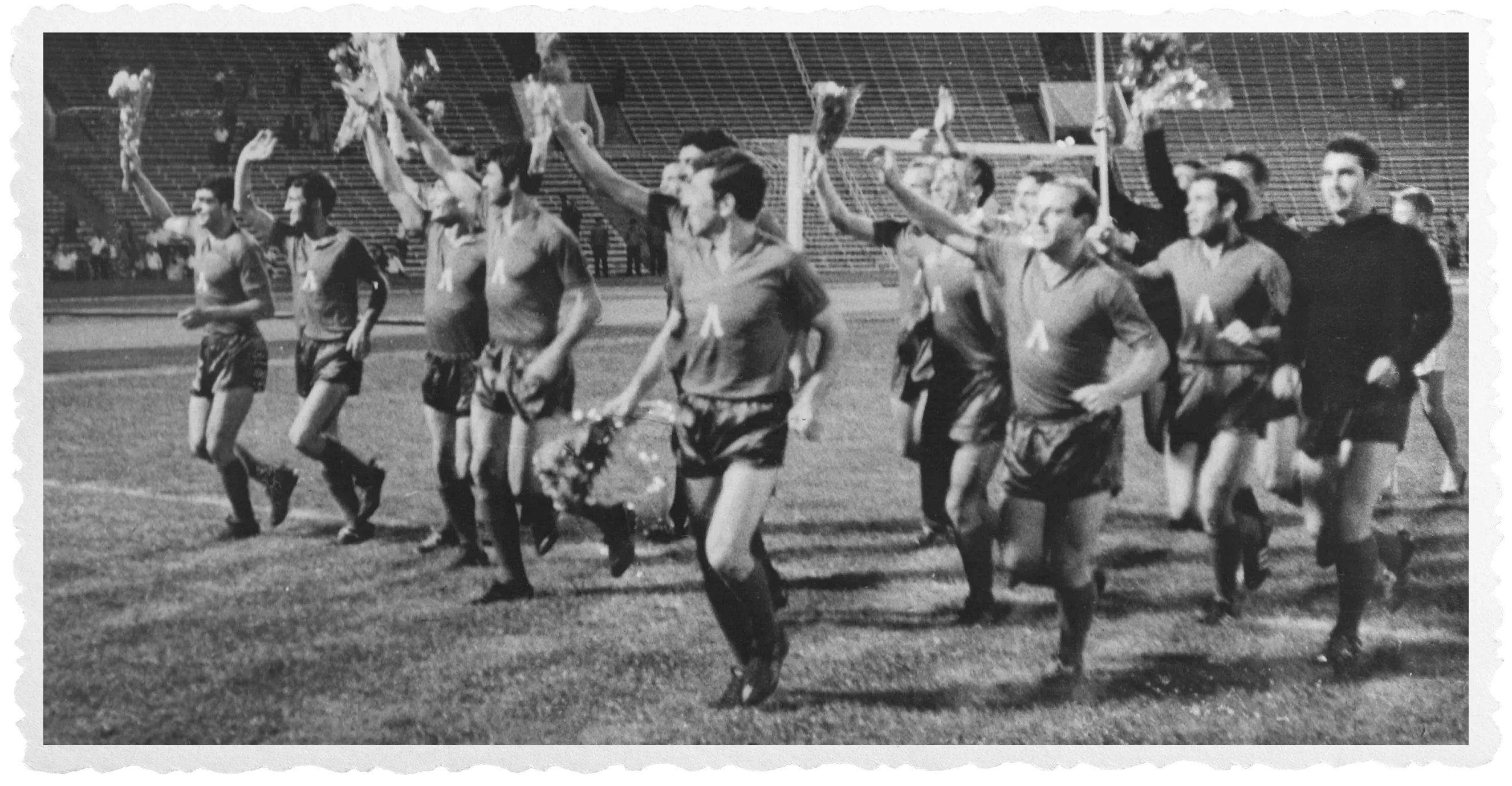 A black-and-white photo of a group of soccer players in sportswear celebrating on a soccer field, some holding bouquets of flowers and raising their arms in joy.