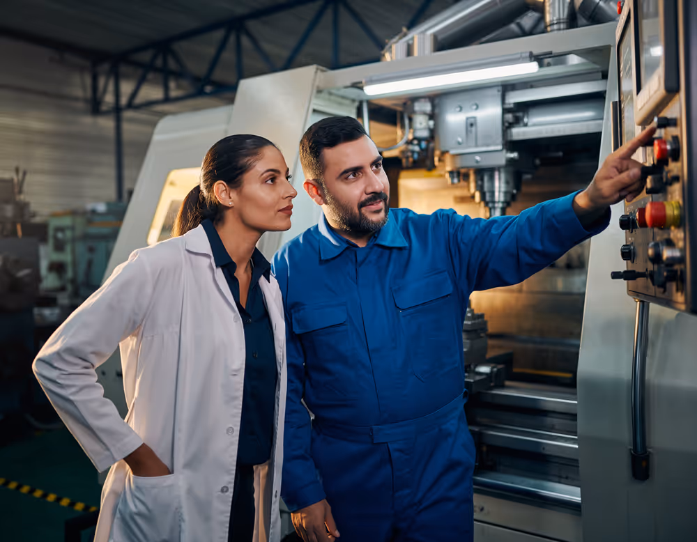Male engineer in blue coveralls operating a control panel on industrial machinery while a female colleague in a white lab coat observes.