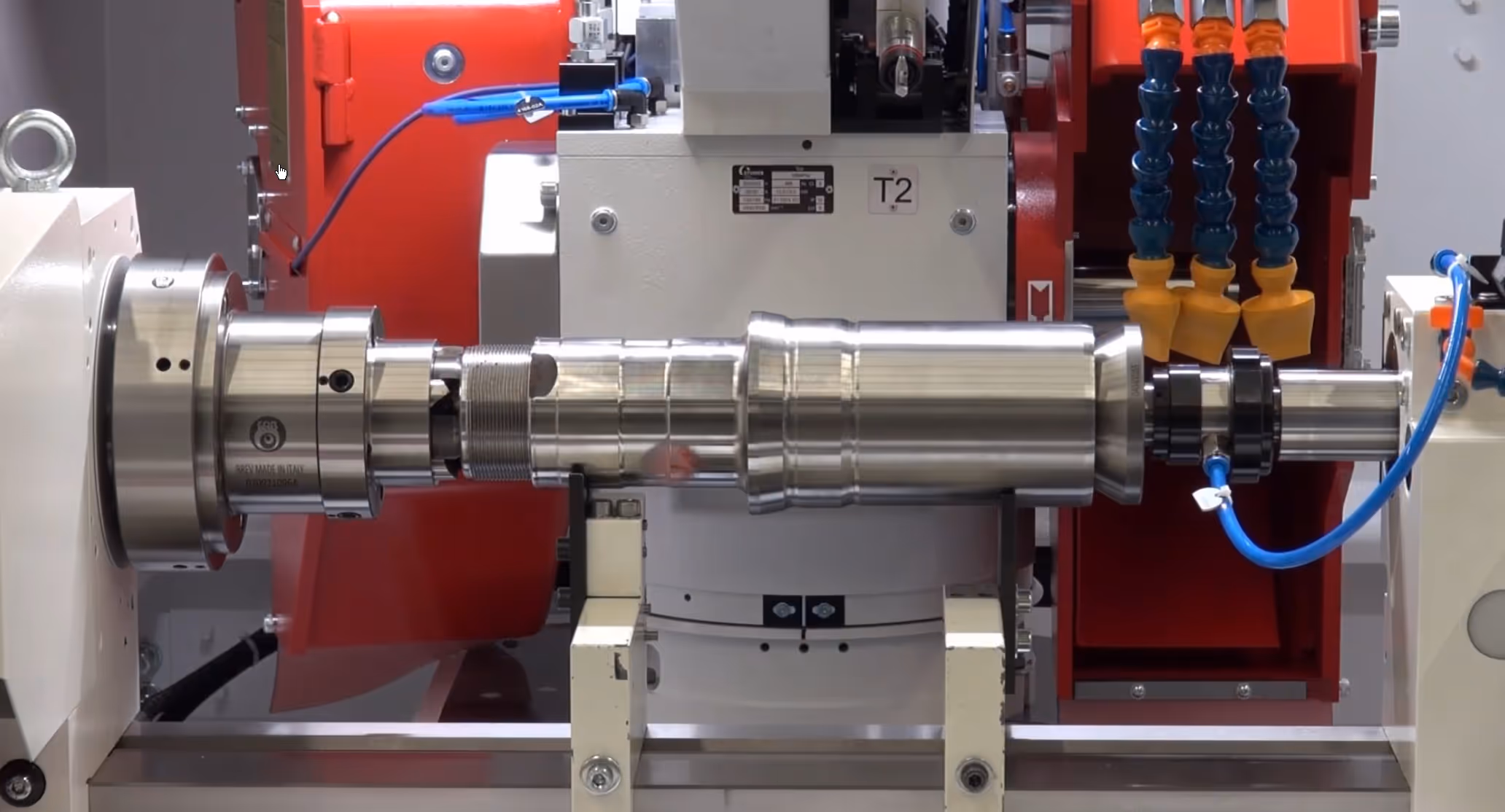 Close-up of a cylindrical metal component mounted on an industrial machining tool with red and white machinery parts and blue tubing.
