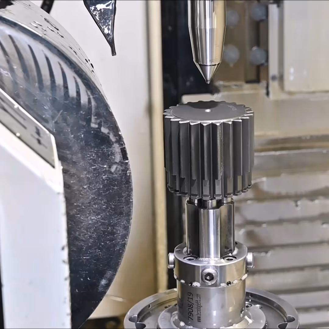 Metal gear mounted on machining equipment, positioned under a pointed polishing or measuring tool.