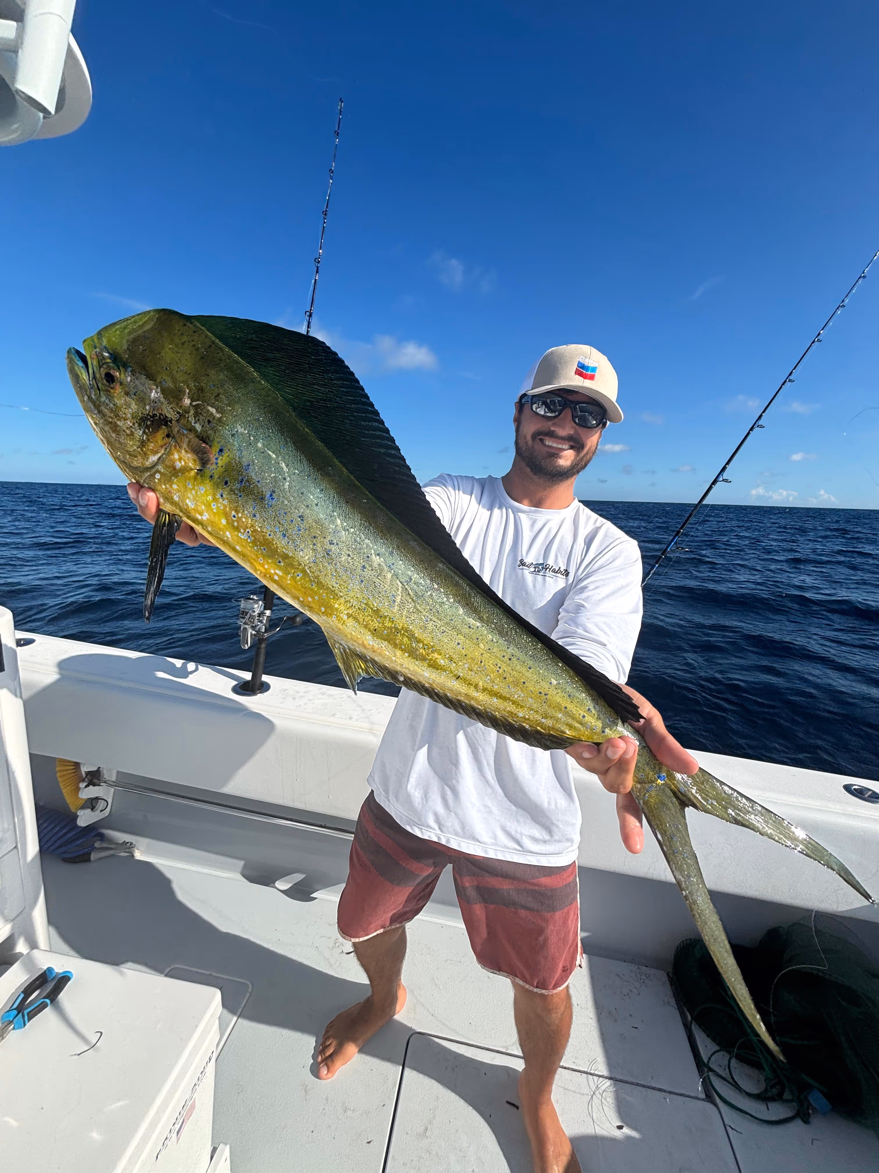 Smiling man wearing sunglasses and a hat holding a large dolphinfish on a boat in the ocean under a clear blue sky.