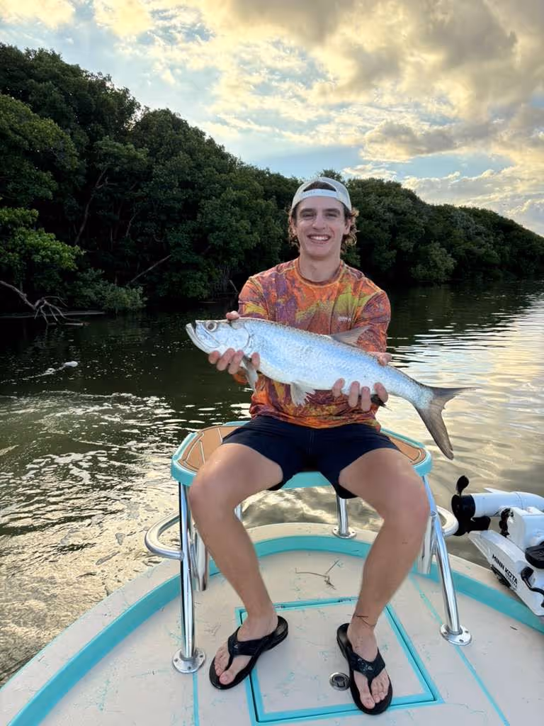 Smiling young man sitting on a boat holding a large silver fish with water and trees in the background.