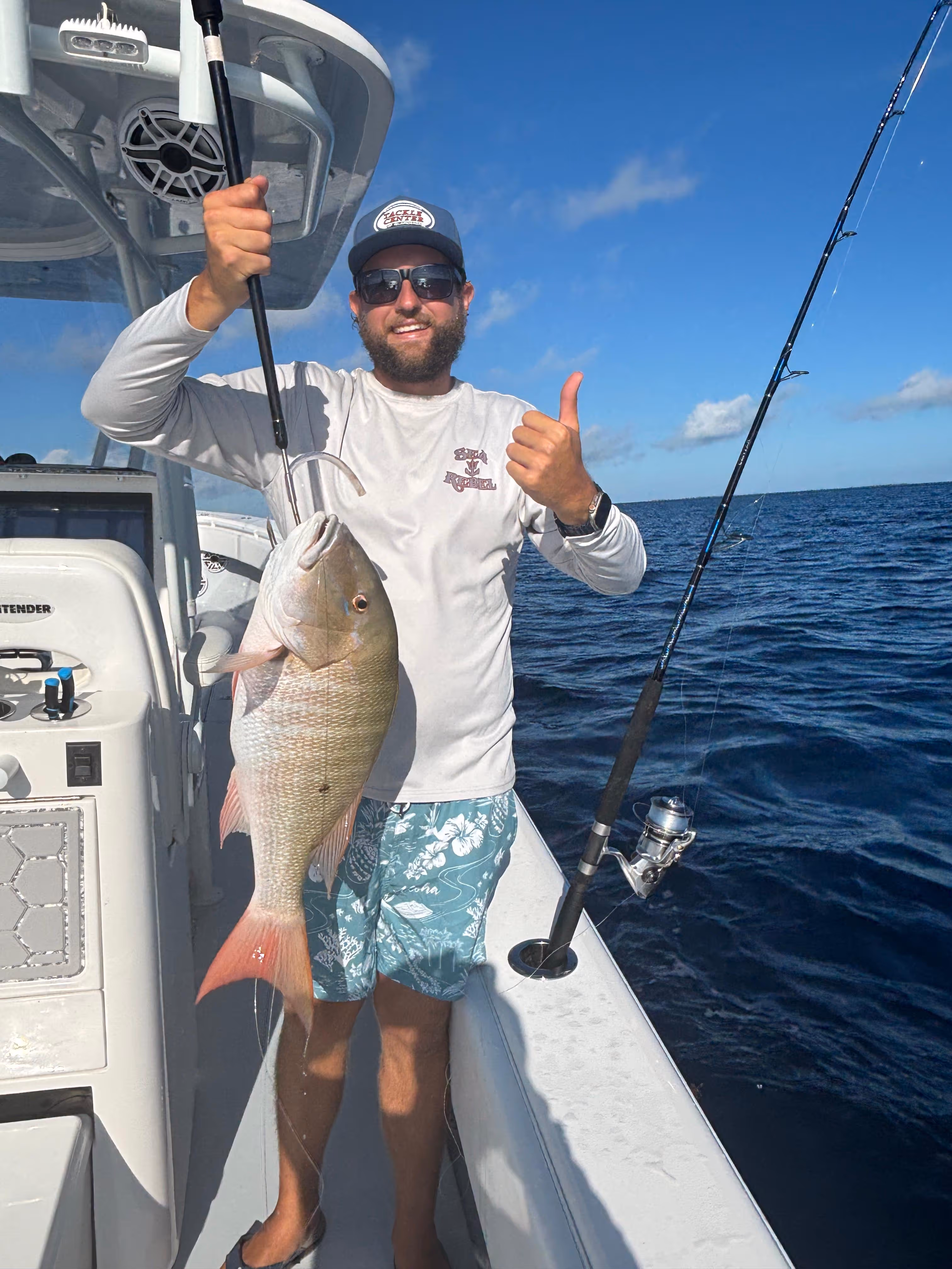 Man wearing sunglasses and a hat on a boat holding a large fish with a fishing rod beside him over open ocean.