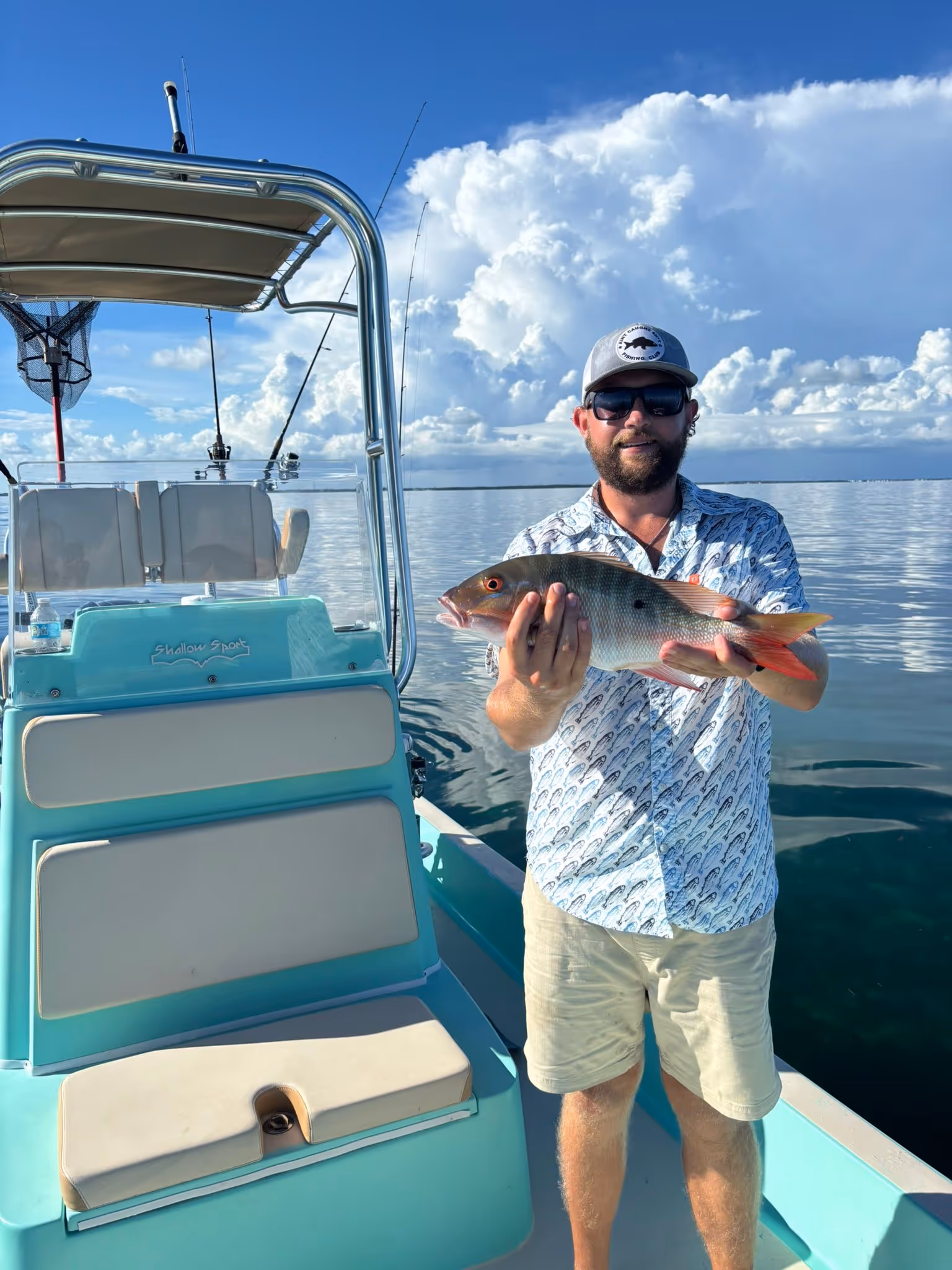 Man wearing sunglasses and a cap holding a redtail snapper fish on a turquoise boat with calm water and cloudy sky in the background.