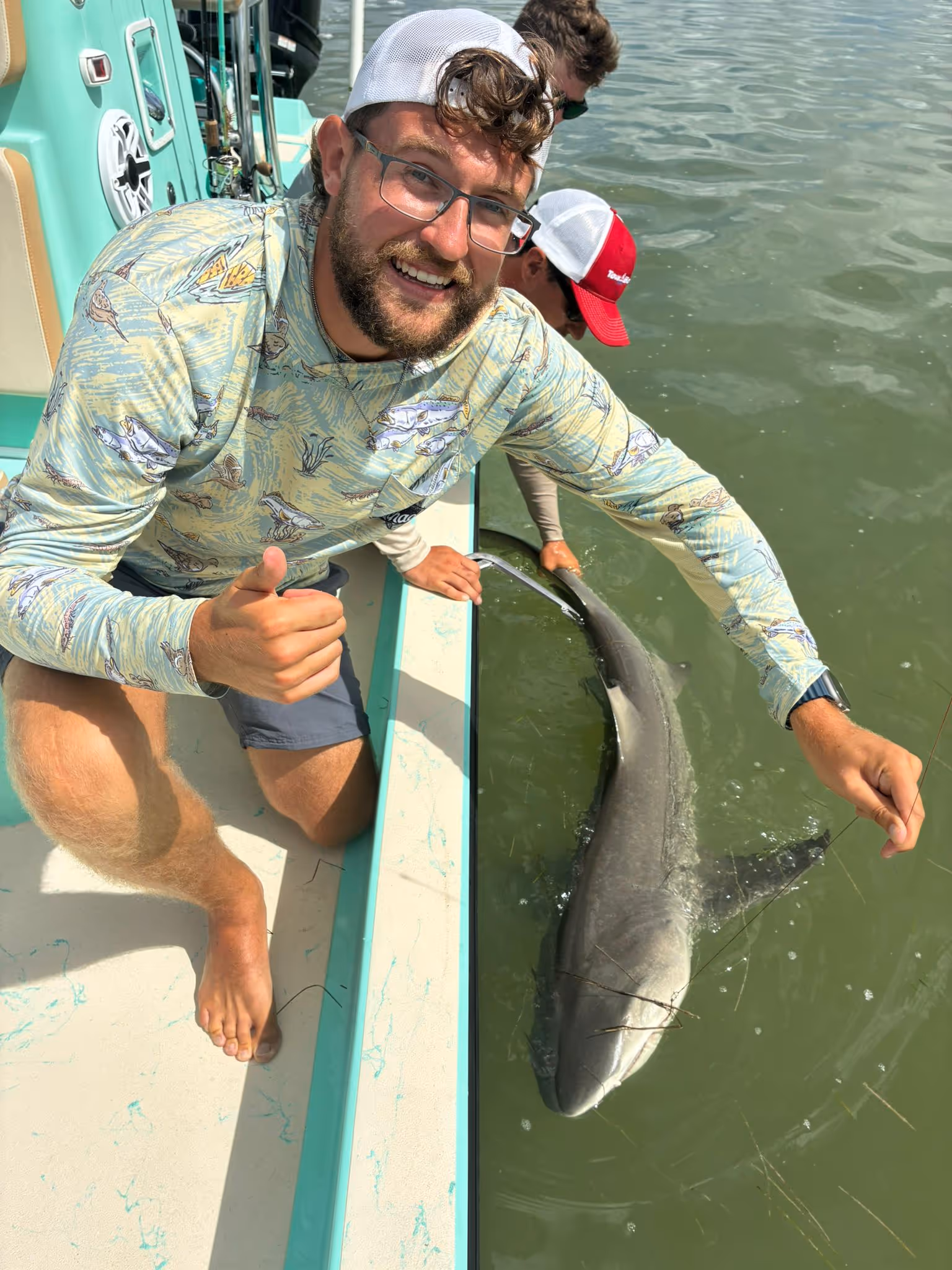 Smiling man giving thumbs up on a boat while holding a shark in the water.