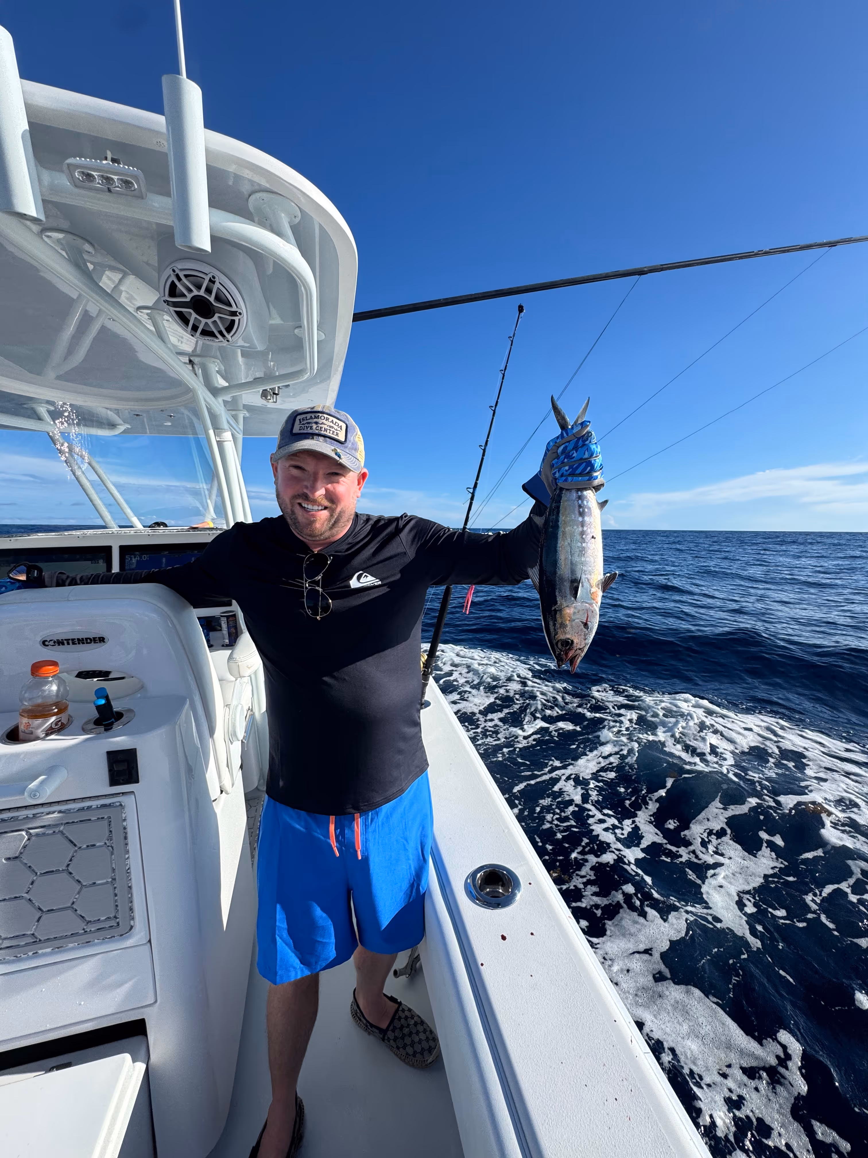 Man smiling and holding a freshly caught fish on a boat in the open ocean under clear blue sky.
