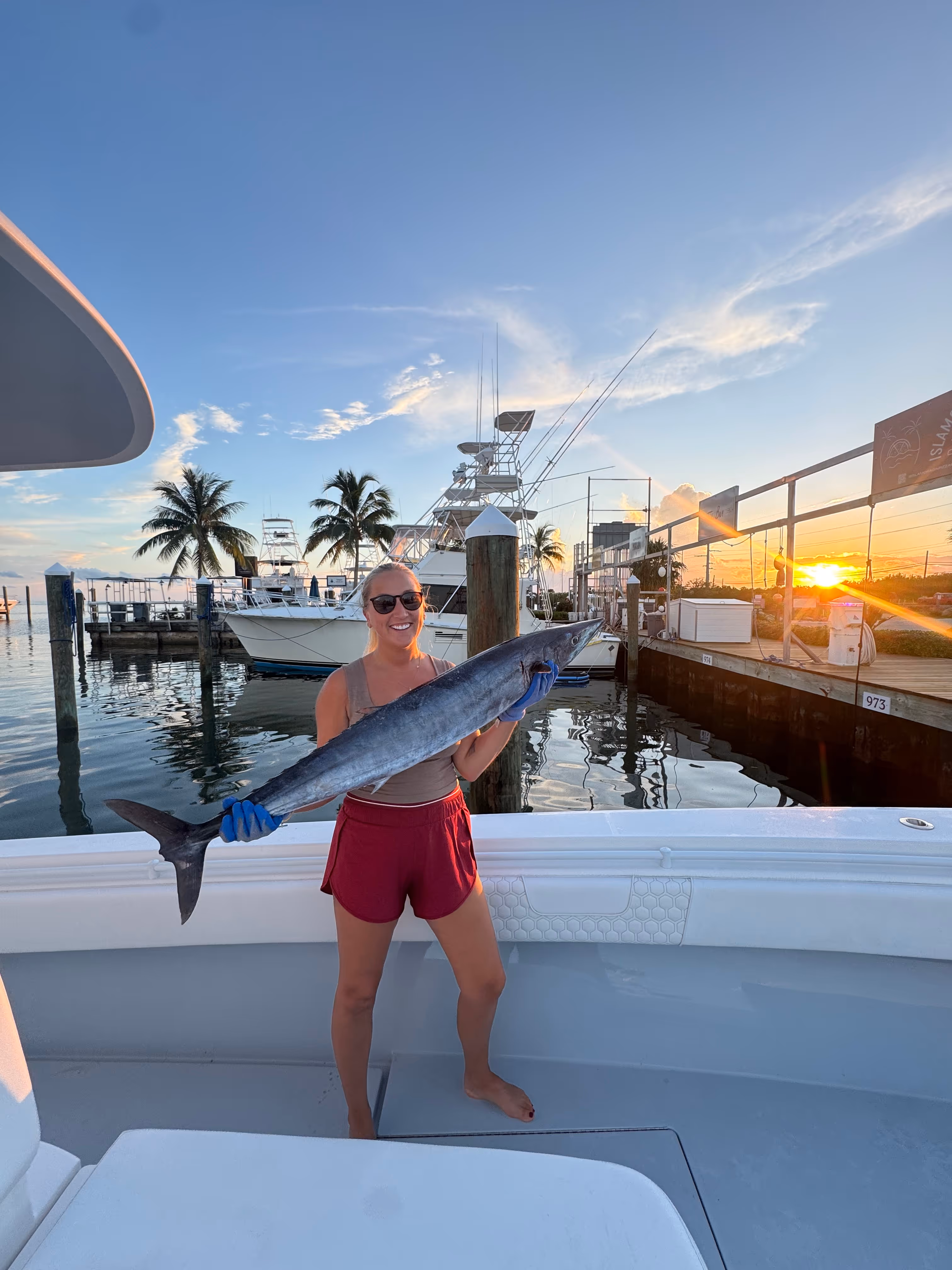 Smiling woman holding a large fish on a boat docked at sunset with palm trees and fishing boats in the background.