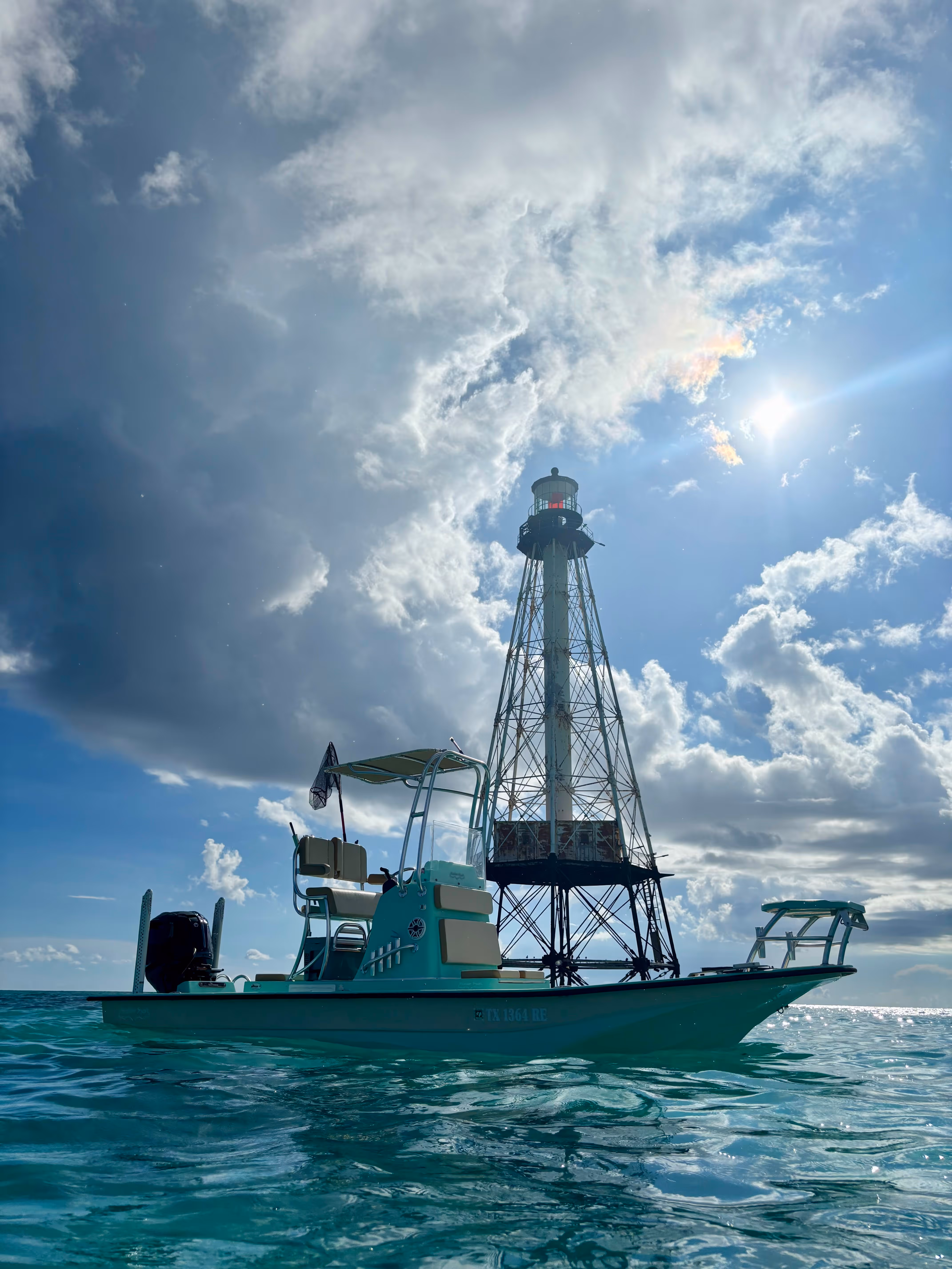 Small boat floating near a tall metal lighthouse under a partly cloudy sky with the sun shining.
