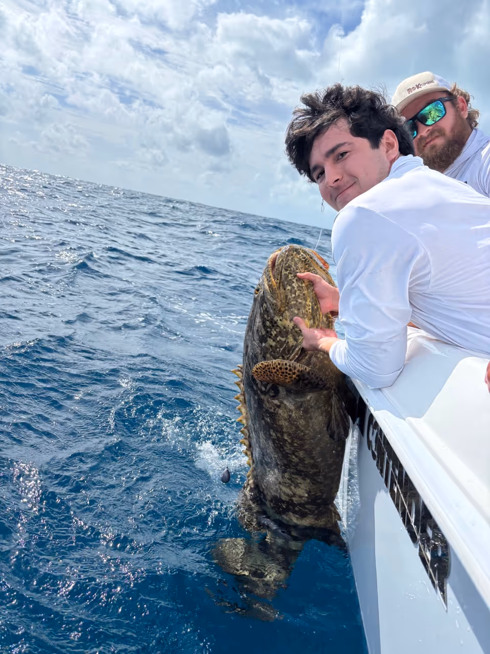 Two men on a boat holding a large grouper fish partially in the water under a cloudy sky.