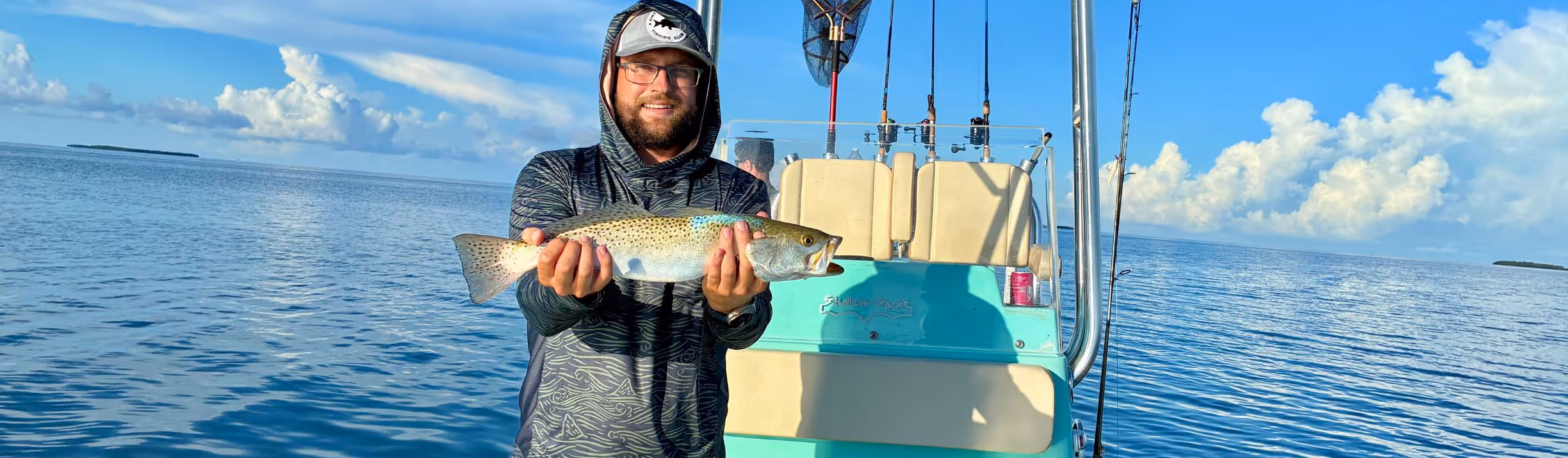 Man wearing hooded shirt and cap holding a spotted fish on a boat with fishing rods and an ocean background under a partly cloudy sky.