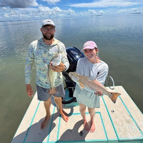 Two people on a boat holding up their catches of fish with calm water and blue sky in the background.