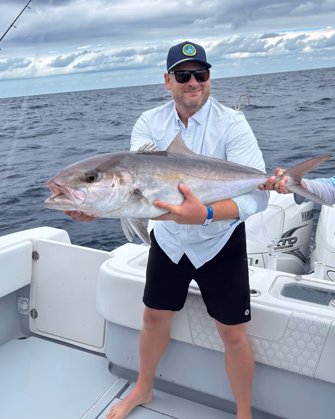 Man wearing sunglasses and a cap holding a large fish on a boat with ocean and cloudy sky in the background.