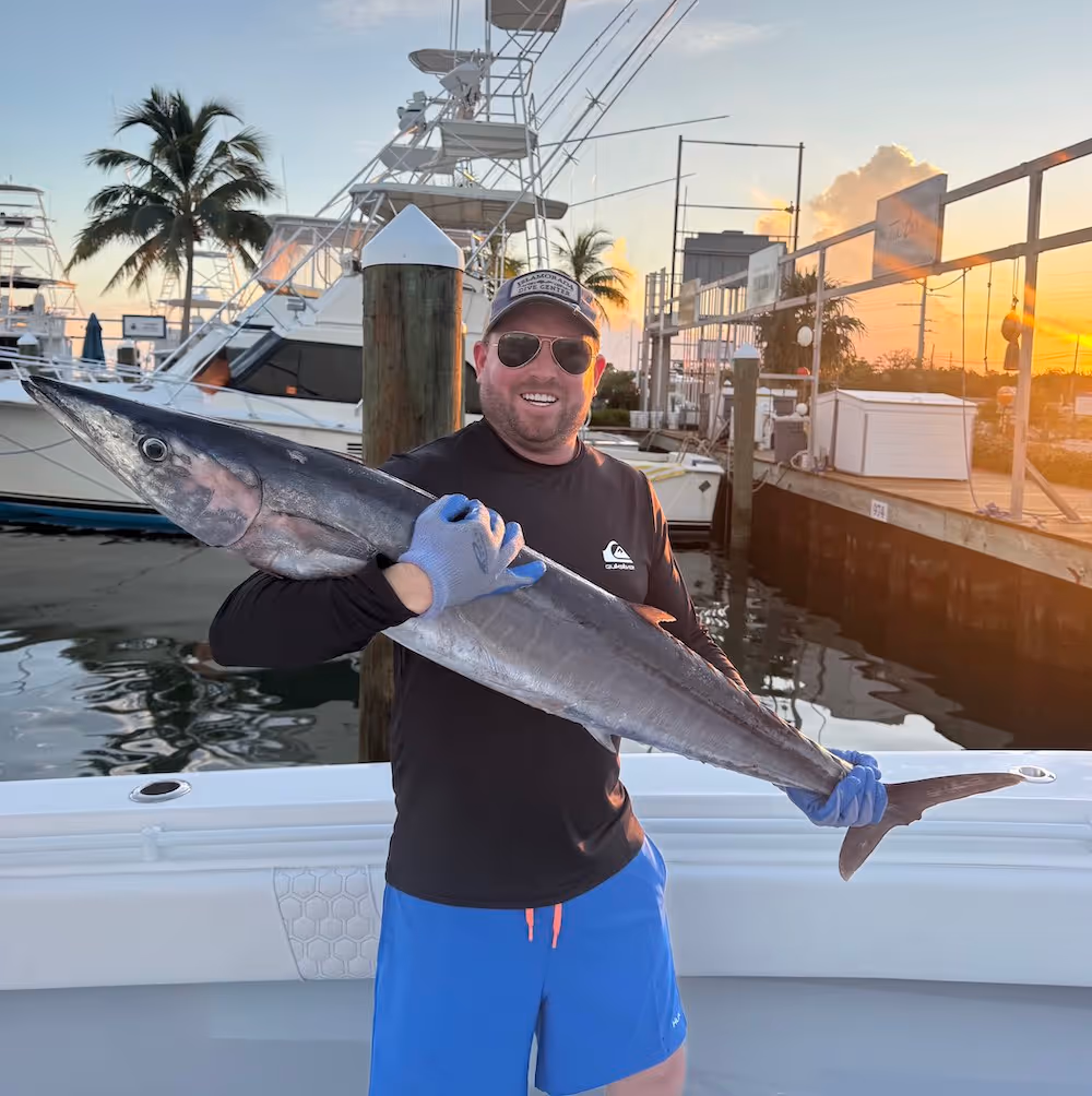 Man wearing sunglasses and gloves holding a large fish on a boat docked at three waters marina with a sunset in the background