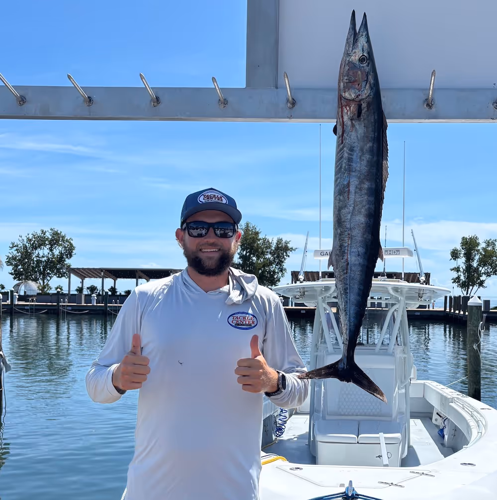 Man wearing sunglasses and a white shirt giving two thumbs up next to a large fish hanging on a boat dock.