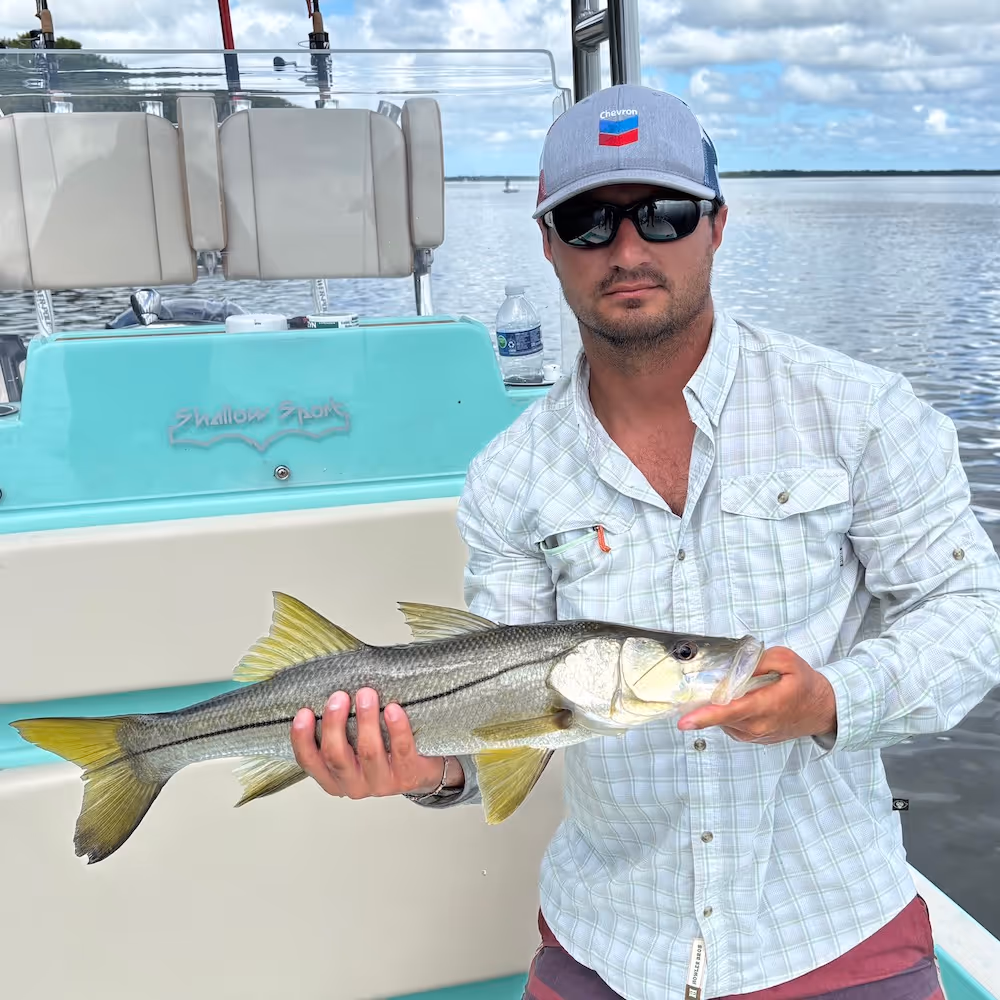 Man wearing sunglasses and a Chevron cap holding a snook on a boat with water and cloudy sky in the background.