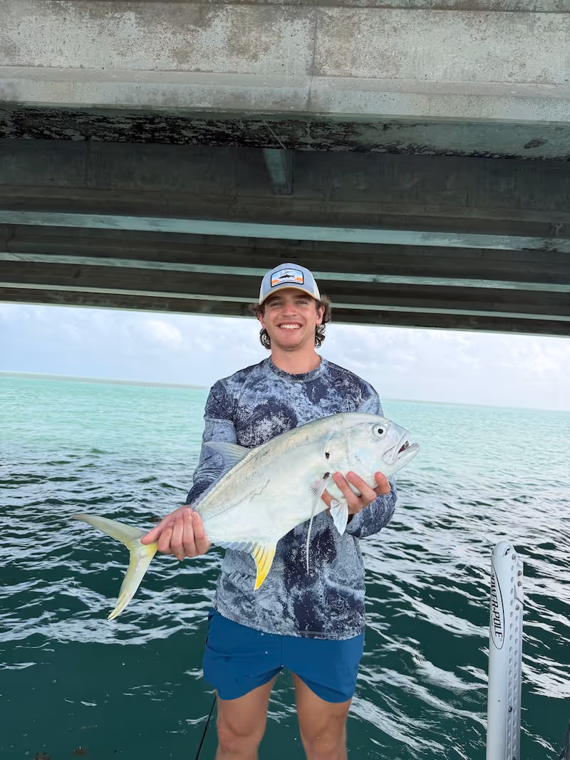 Smiling young man wearing a cap and camouflage shirt holding a large permit on a boat under a bridge with turquoise water in the background.