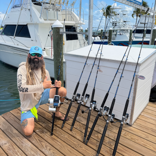Captain Peter Bernal, who guides fishing tours in Islamorada, posing next to a line of fishing rods near the water. A large cooler and a white fishing boat are behind him in the background.
