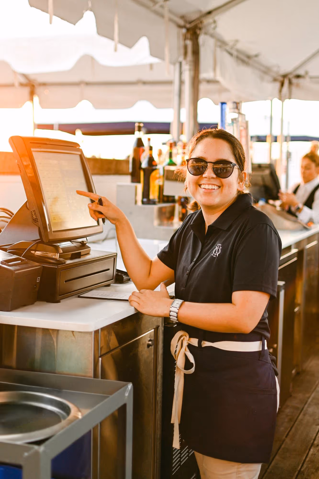 Smiling female bartender wearing sunglasses points at a touchscreen register in a bright bar setting.
