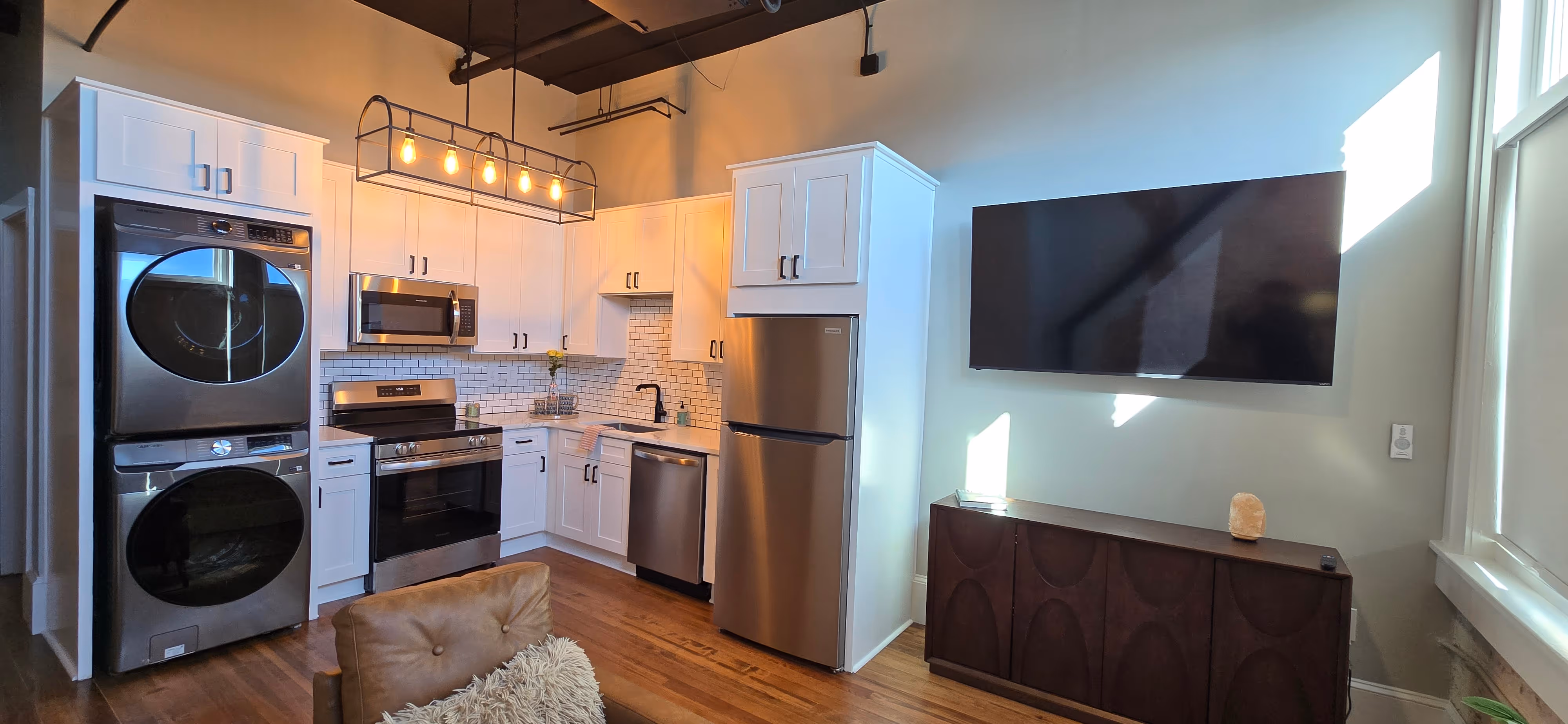 Modern kitchen with white cabinets, stainless steel appliances including stacked washer and dryer, stove, microwave, dishwasher, and refrigerator, a hanging light fixture, wooden floor, and a mounted flat screen TV above a dark wooden cabinet.