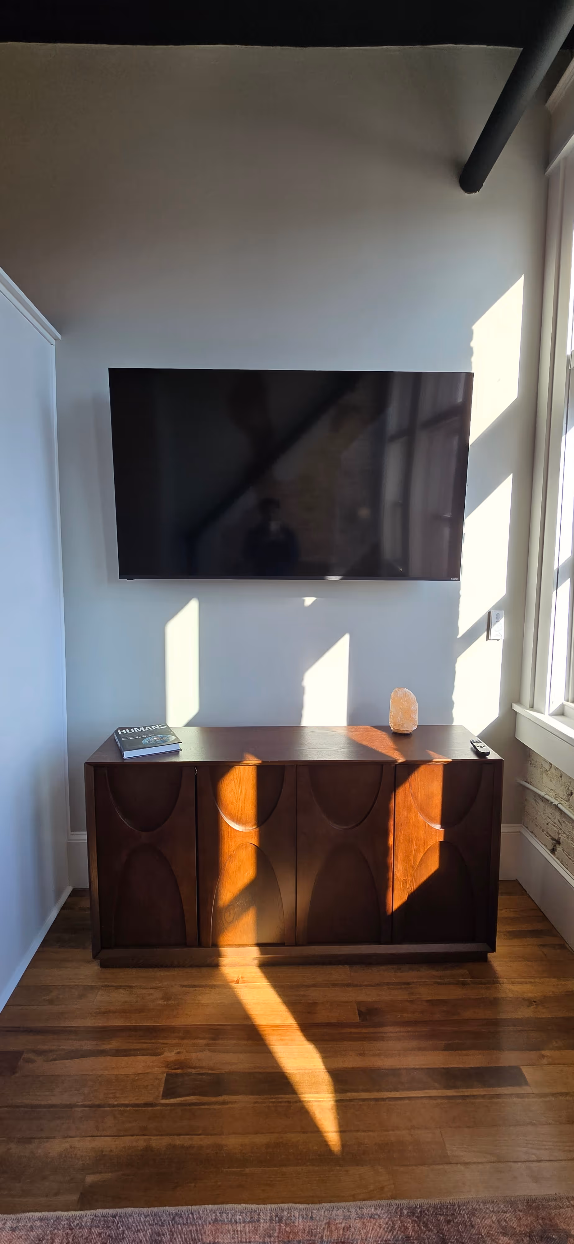 Flat-screen TV mounted on white wall above wooden cabinet with a book, salt lamp, and remote, sunlight casting shadows.