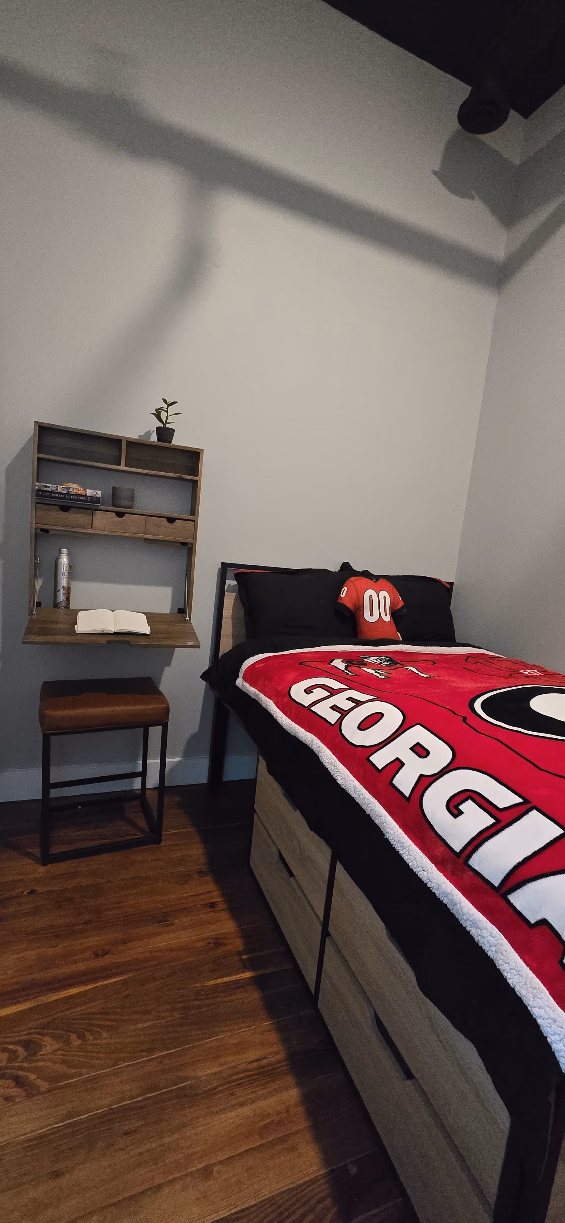 Small bedroom corner with a wooden bed covered by a red Georgia Bulldogs blanket and a matching jersey pillow, a wall-mounted wooden desk with a plant, books, a cup, and an open notebook, and a brown stool on a wooden floor.