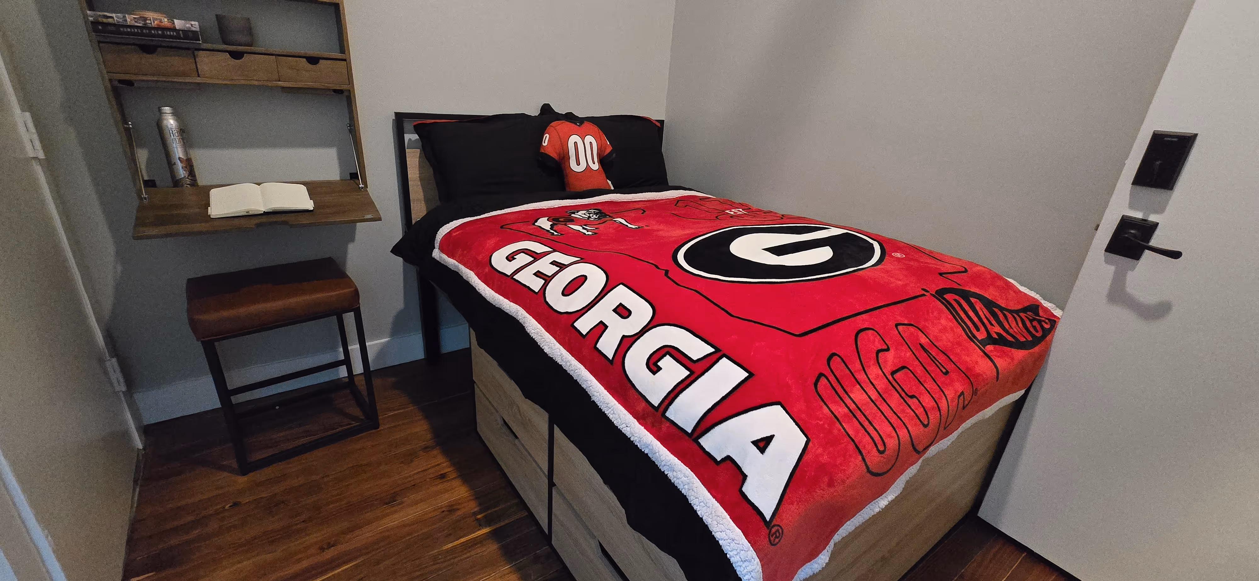 Bedroom with a bed featuring a red University of Georgia blanket and black pillows, one with a red jersey displaying number 00, next to a small desk with an open book and a stool.