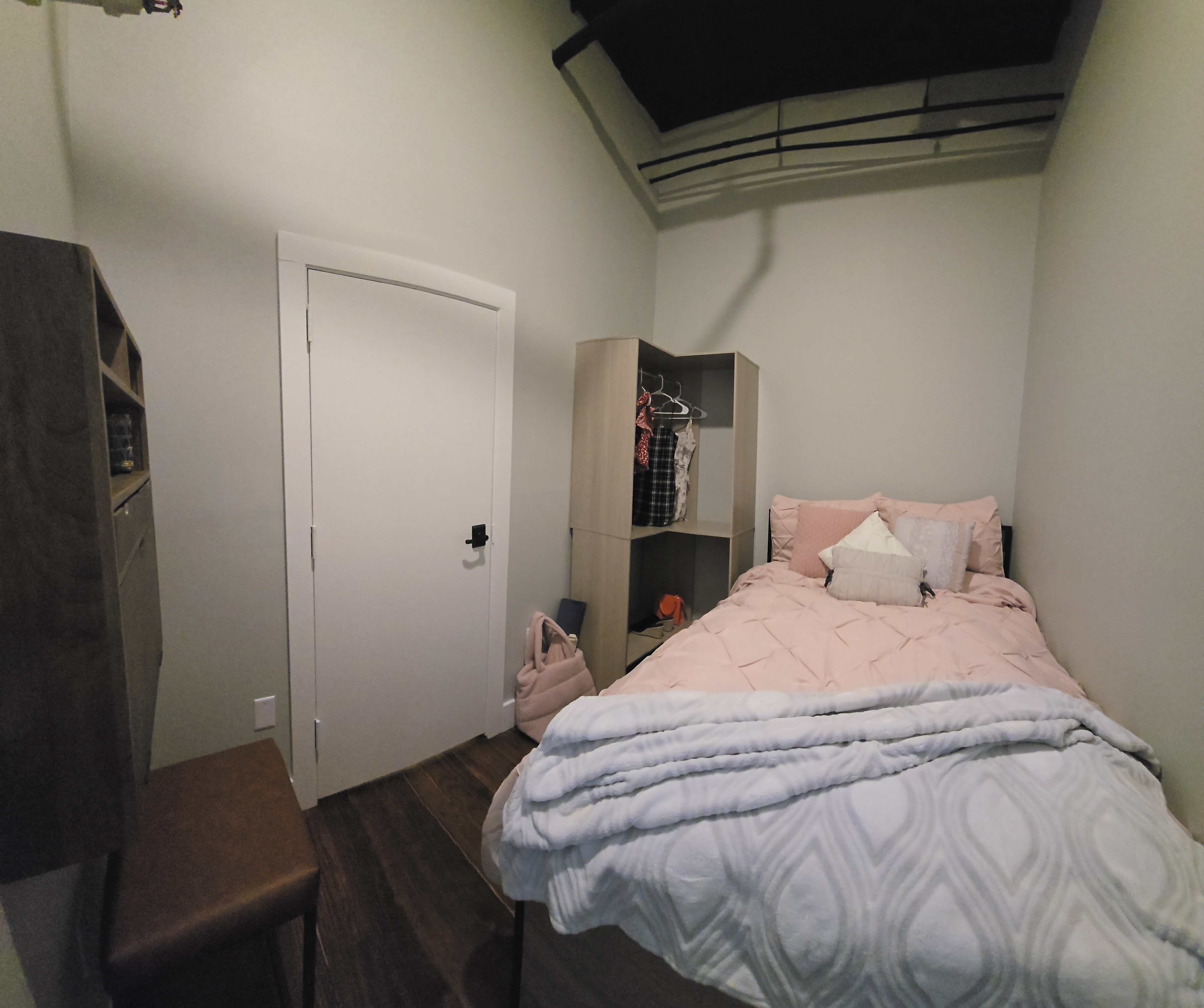 Small bedroom with a bed made with pink bedding and several pillows, a white blanket folded at the foot, a wooden shelf with hanging clothes, and a closed white door on the left wall.