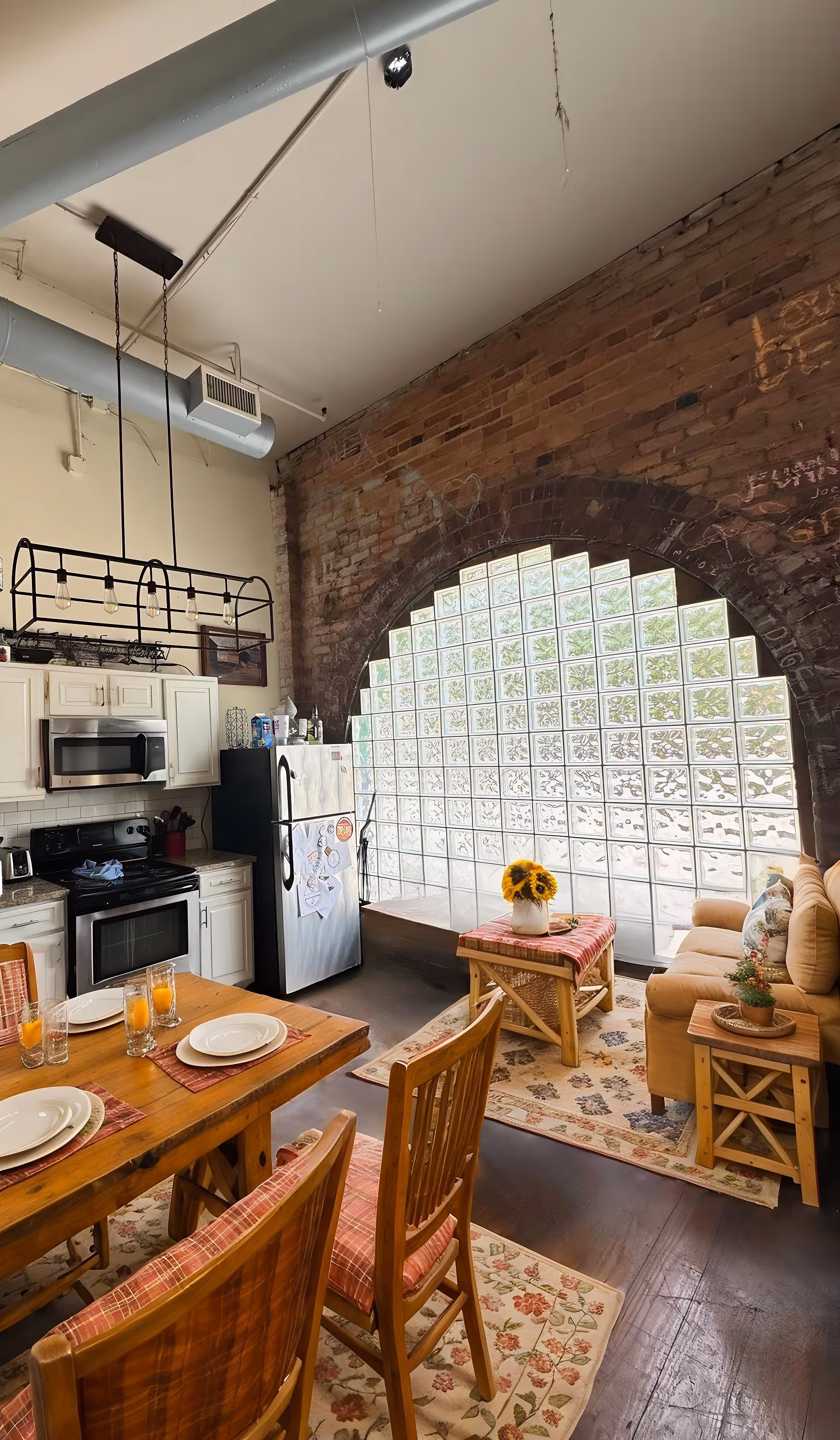 Cozy kitchen and living area with wooden dining table, chairs, beige sofa, glass block arched window, and brick wall.