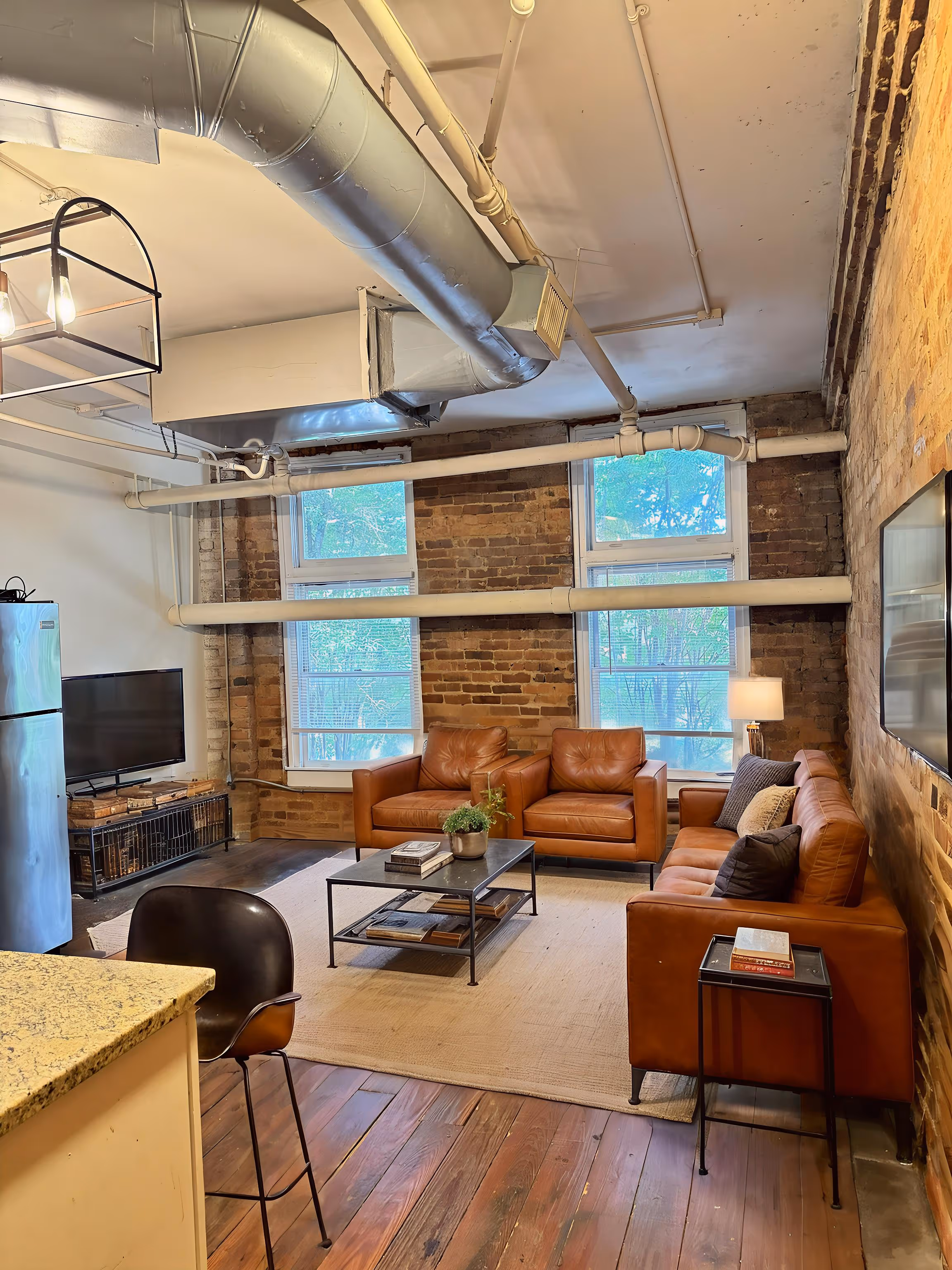 Cozy living room with brown leather sofa and armchairs, exposed brick walls, large windows, and a flat-screen TV.