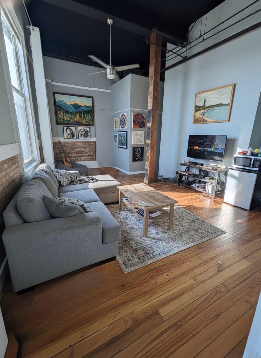 Sunlit living room with a gray sectional sofa, wooden coffee table on a patterned rug, wall art, a TV on a stand, a fan, and hardwood floors.