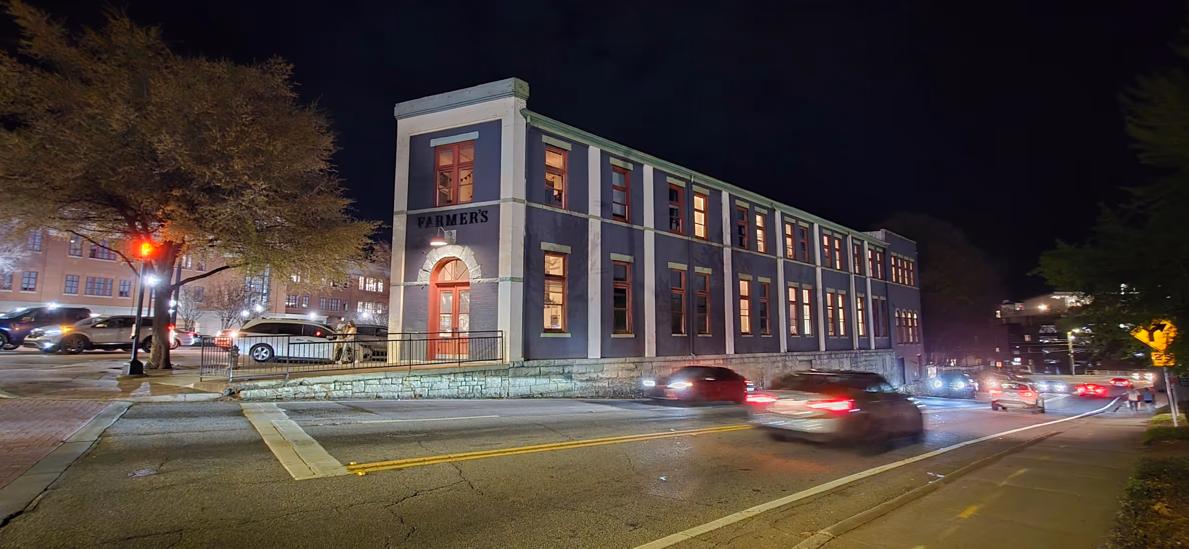Night view of a two-story building with 'FARMER'S' sign, lit windows, and cars passing on the street.