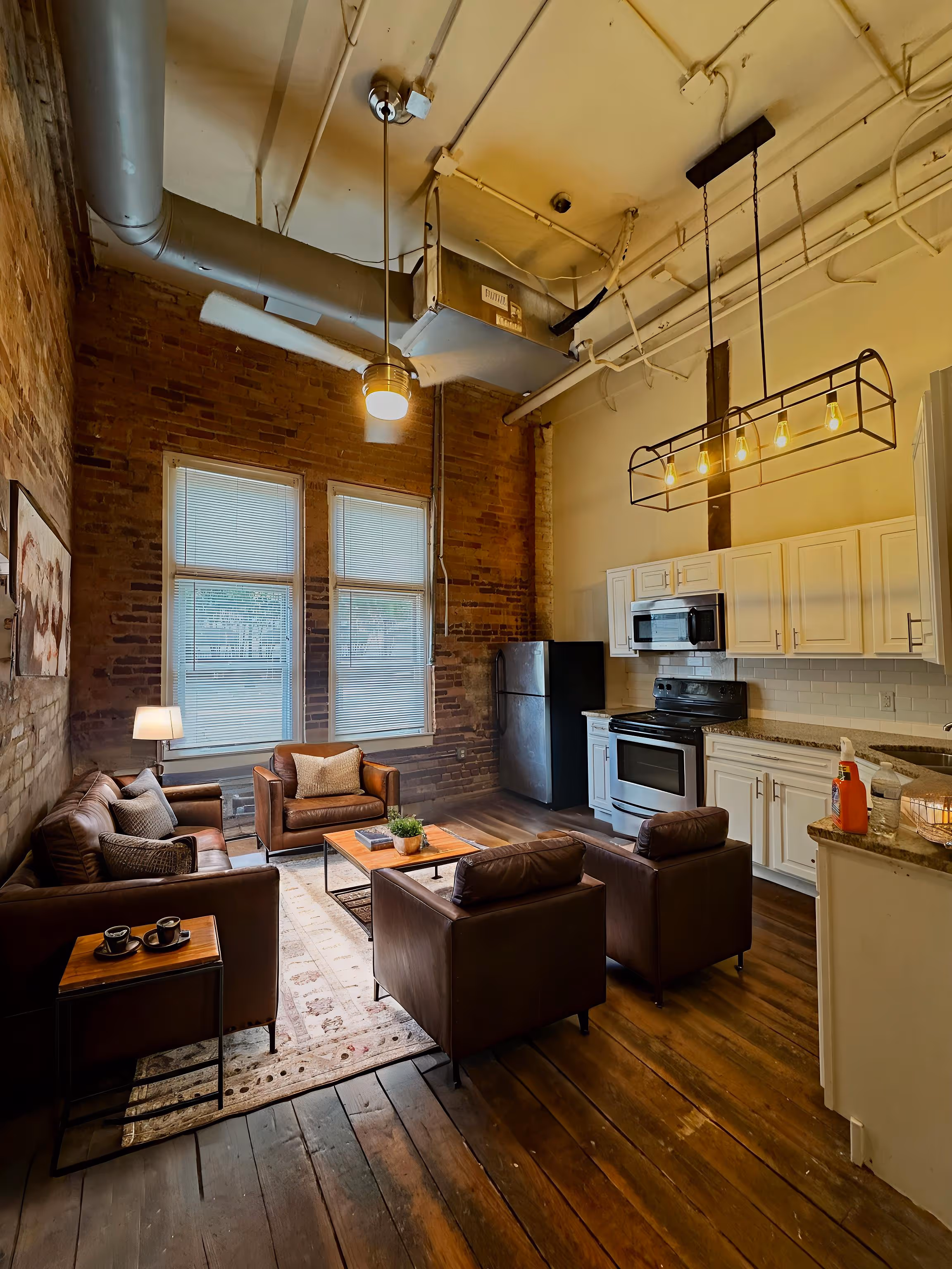 Cozy loft-style living room and kitchen with exposed brick walls, wooden floor, leather sofas, and modern kitchen appliances.