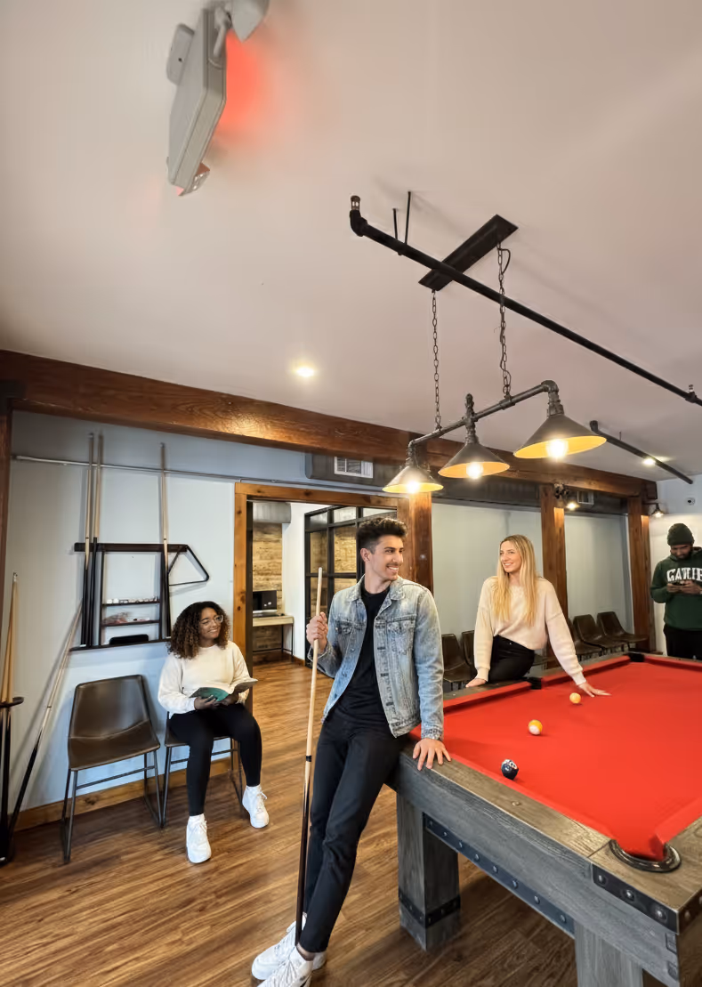 Three people in a game room with a red pool table, one holding a cue stick, one sitting on the table, and one seated reading a book.