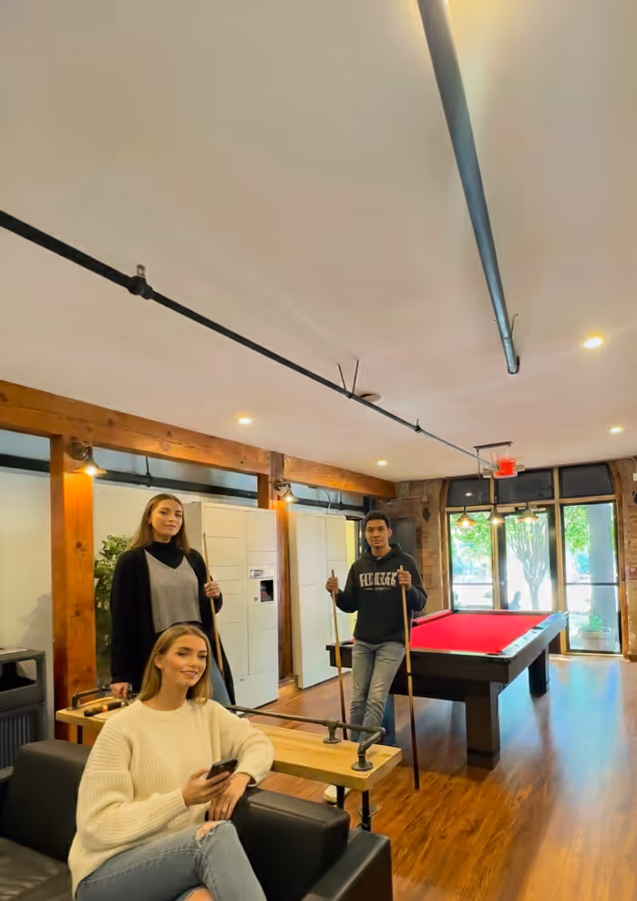 Three young adults in a modern game room with a red pool table; one woman is seated on a black couch holding a phone, while a man and another woman stand holding pool cues.