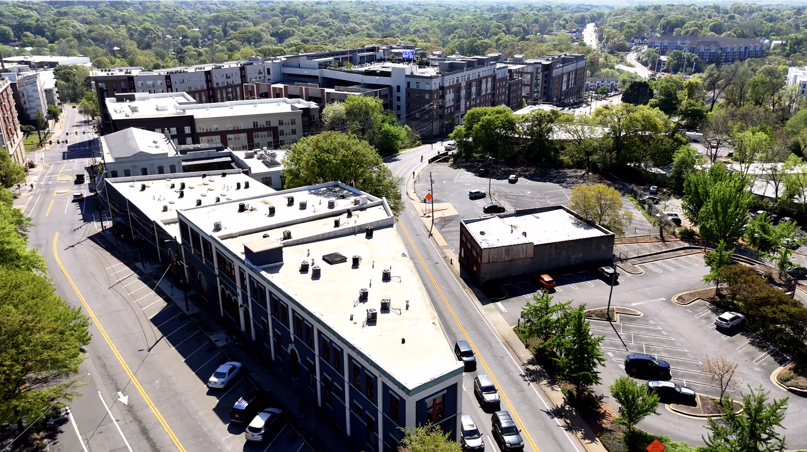 Aerial view of a city block with multi-story buildings, parking lots, and surrounding green trees on a sunny day.