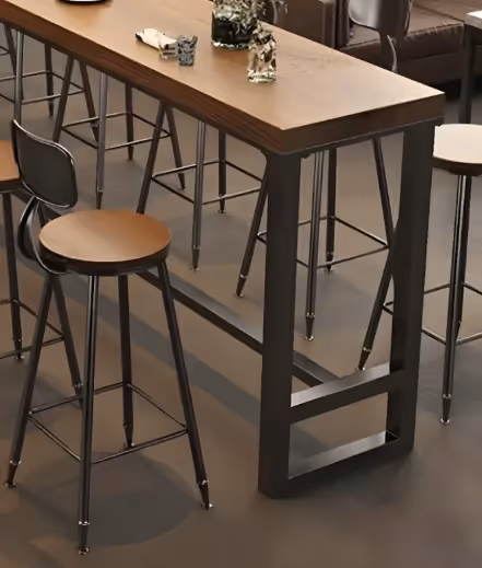 Modern high wooden table with black metal legs and matching round wooden stools in a room with brown flooring and dark furniture.