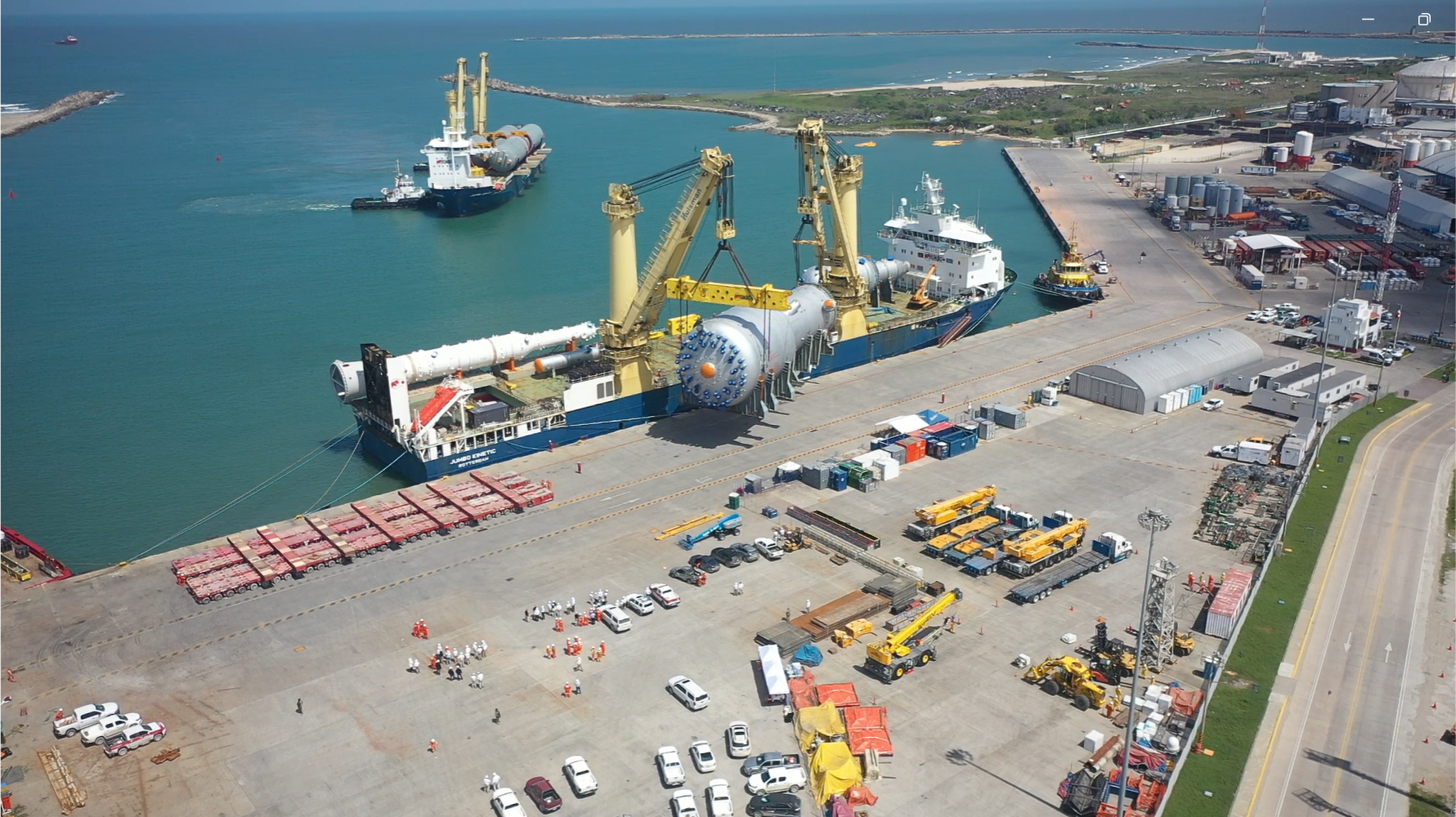 Aerial view of a busy port with two cargo ships docked; one ship is using cranes to load or unload a large industrial vessel part while workers and vehicles are dispersed across the concrete dock area.