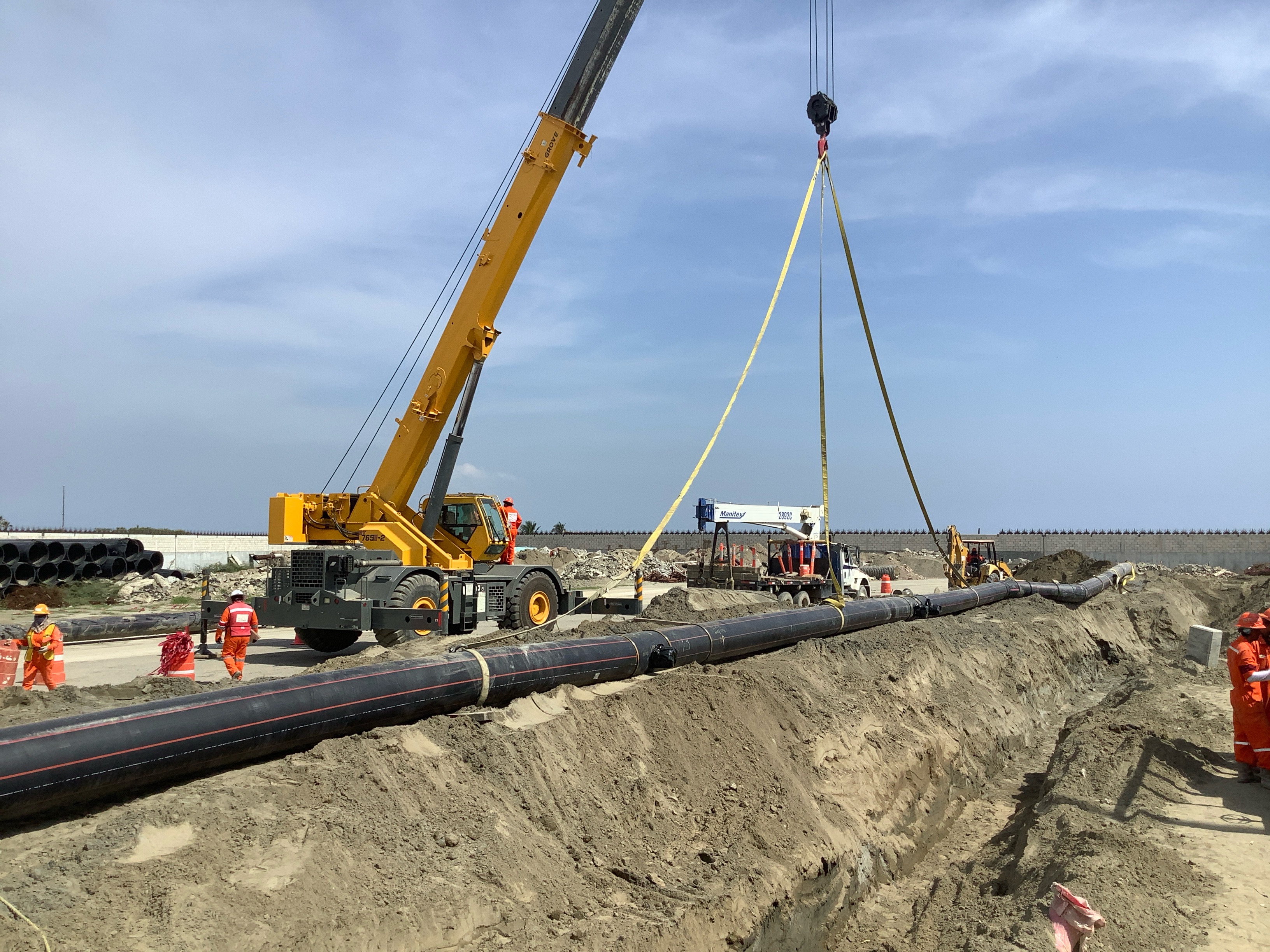 Construction site with workers in orange safety gear installing a large pipeline lifted by a yellow crane over a dirt trench.