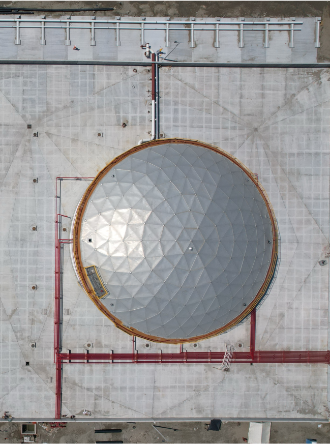 Aerial view of a large circular industrial storage tank with geometric patterned metal roof and surrounding pipes on a concrete surface.