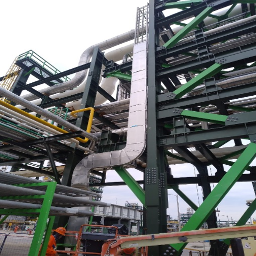 Industrial facility with large metal pipes, green and black steel framework, and workers in orange safety gear.