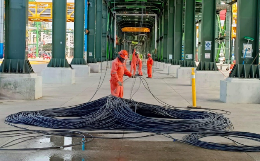 Workers in orange safety suits handling large coiled cables under a green metal structure in an industrial setting.