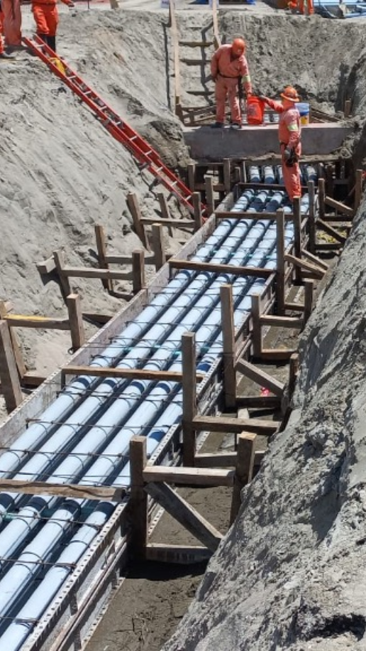Construction workers in orange safety gear working on installing multiple large blue pipes in a deep trench with wooden support structures and ladders.
