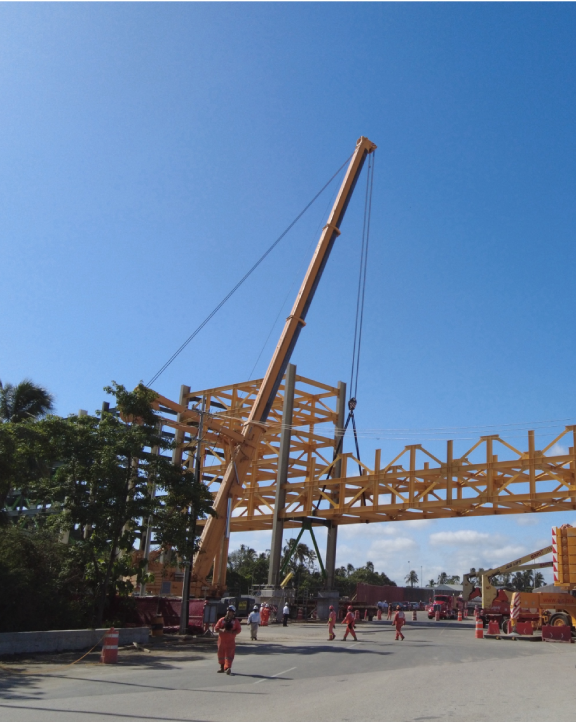 Construction workers in orange uniforms operating a large yellow crane lifting a steel structure on a clear day.